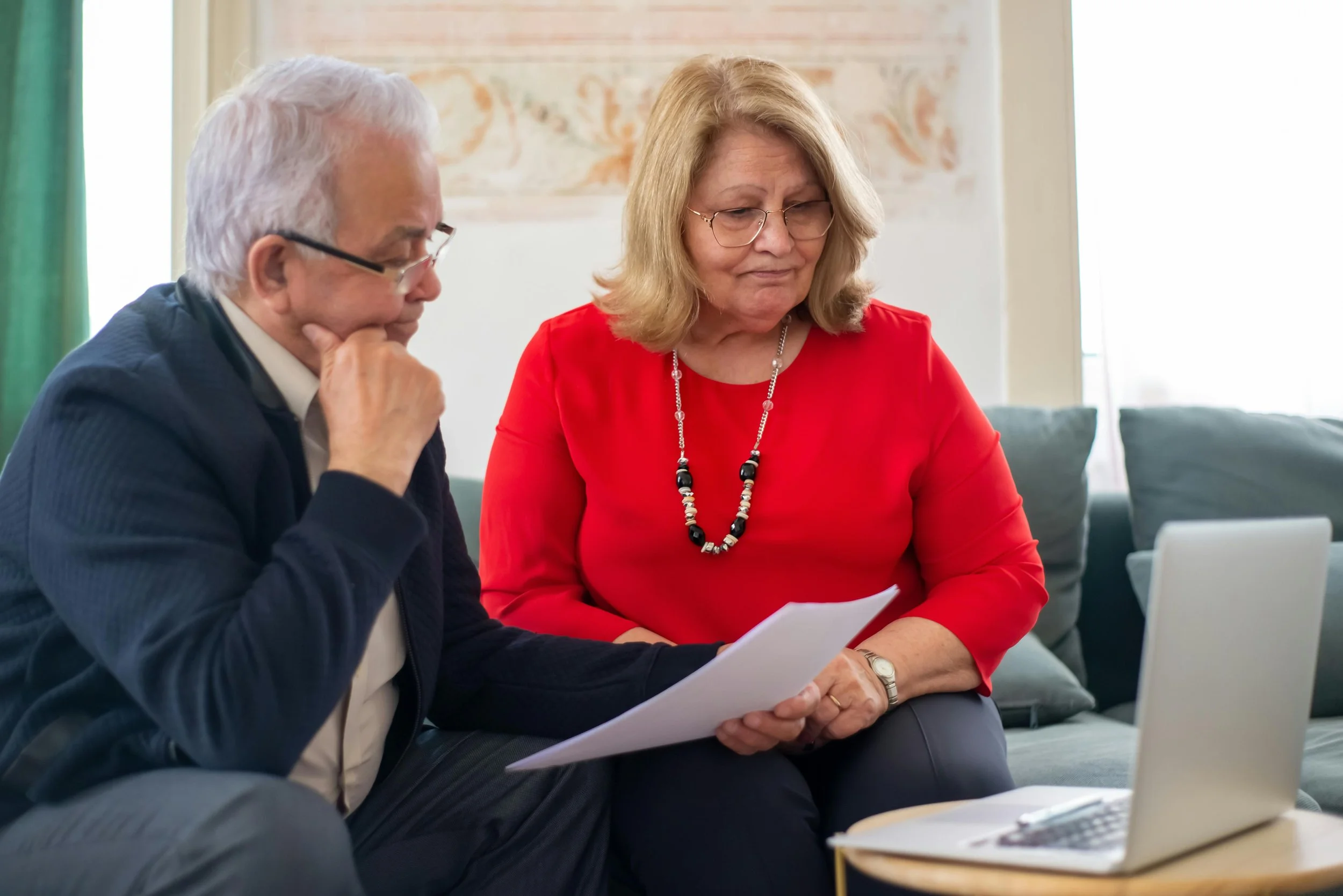 An older man and woman sitting on a couch looking at papers and a laptop in a cozy living room.