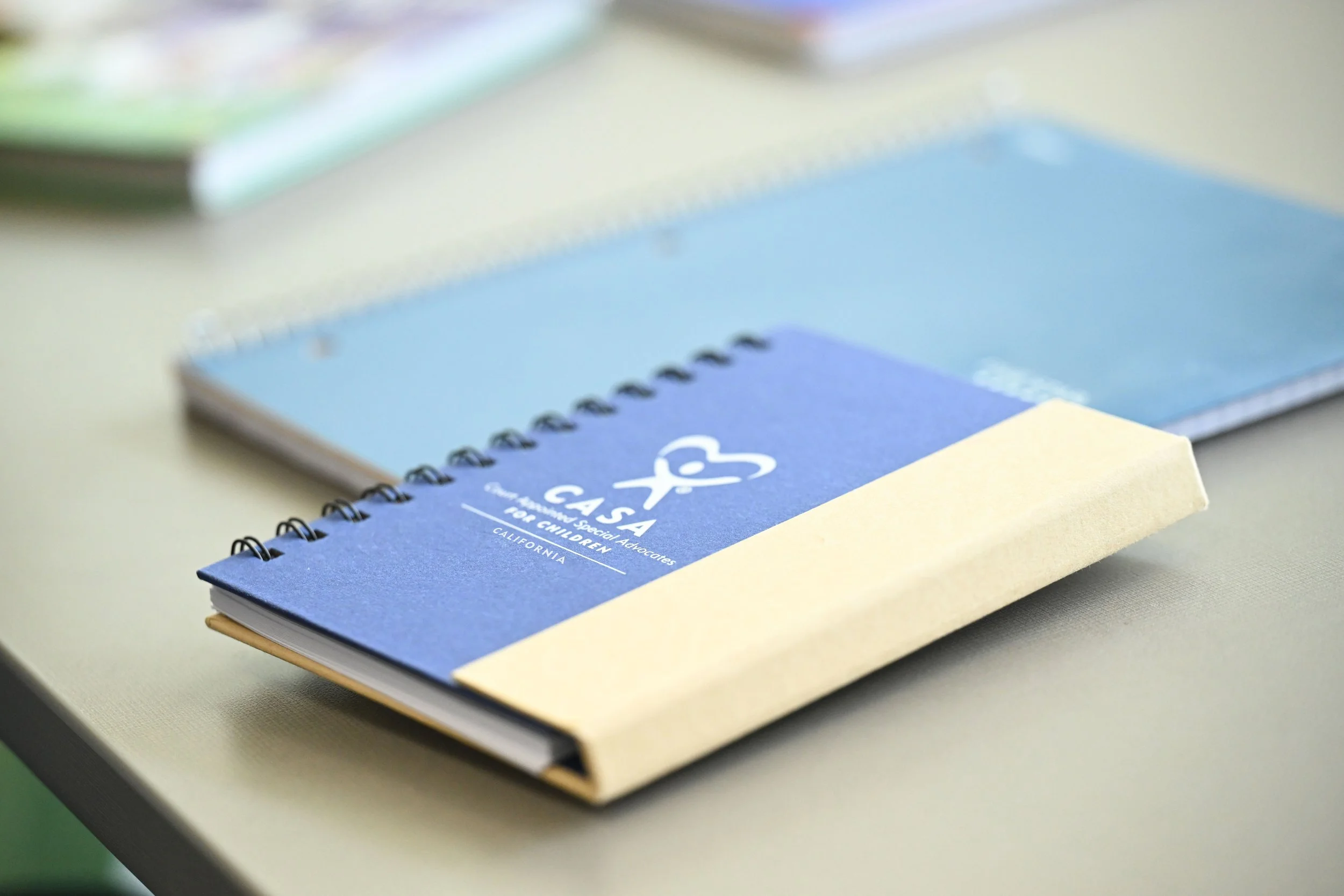 A blue and tan spiral-bound notebook labeled Casa for Children, California, resting on a light-colored table with other notebooks blurred in the background.