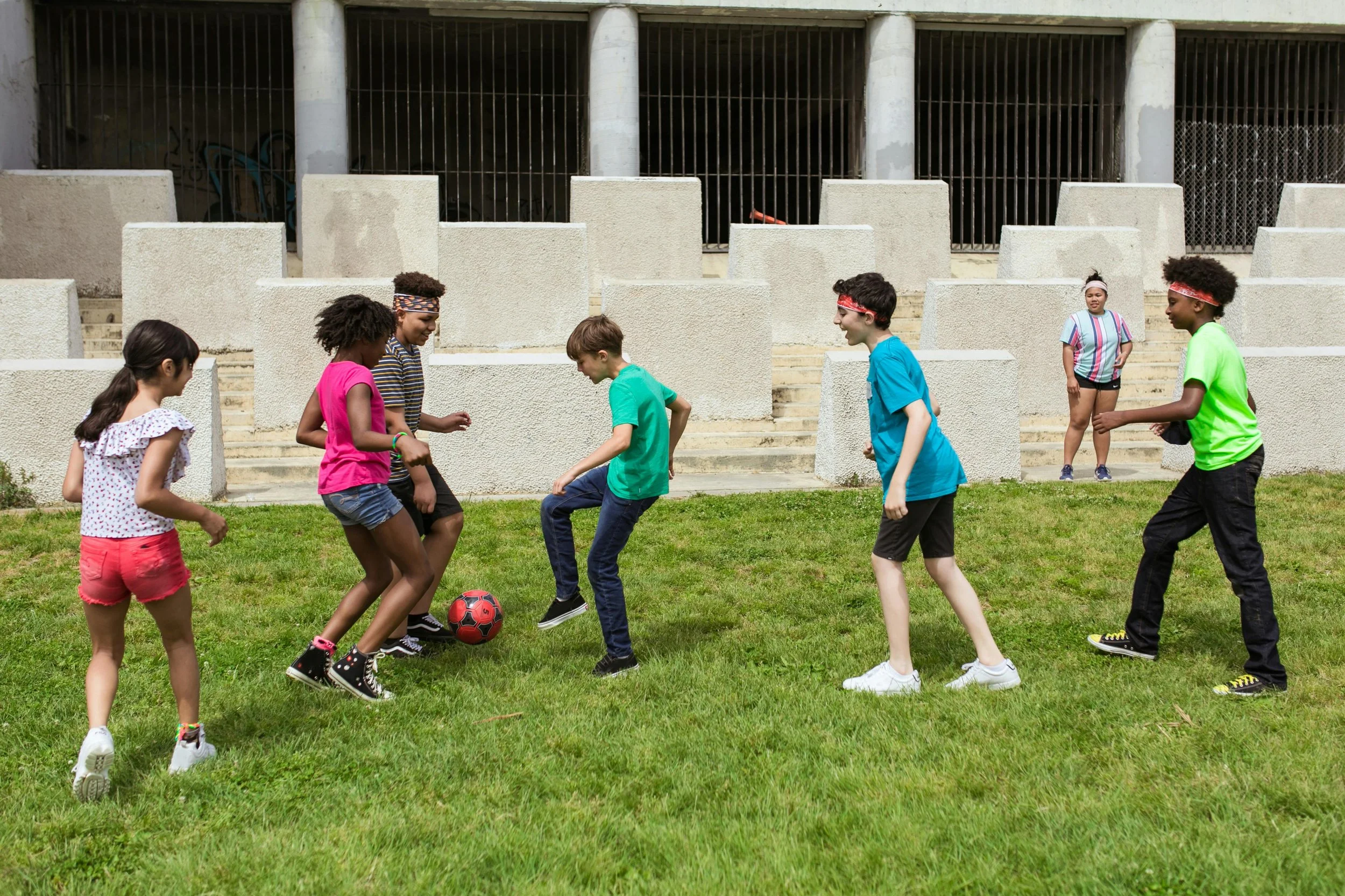 Children playing soccer on a grassy field with a concrete structure and building behind them.