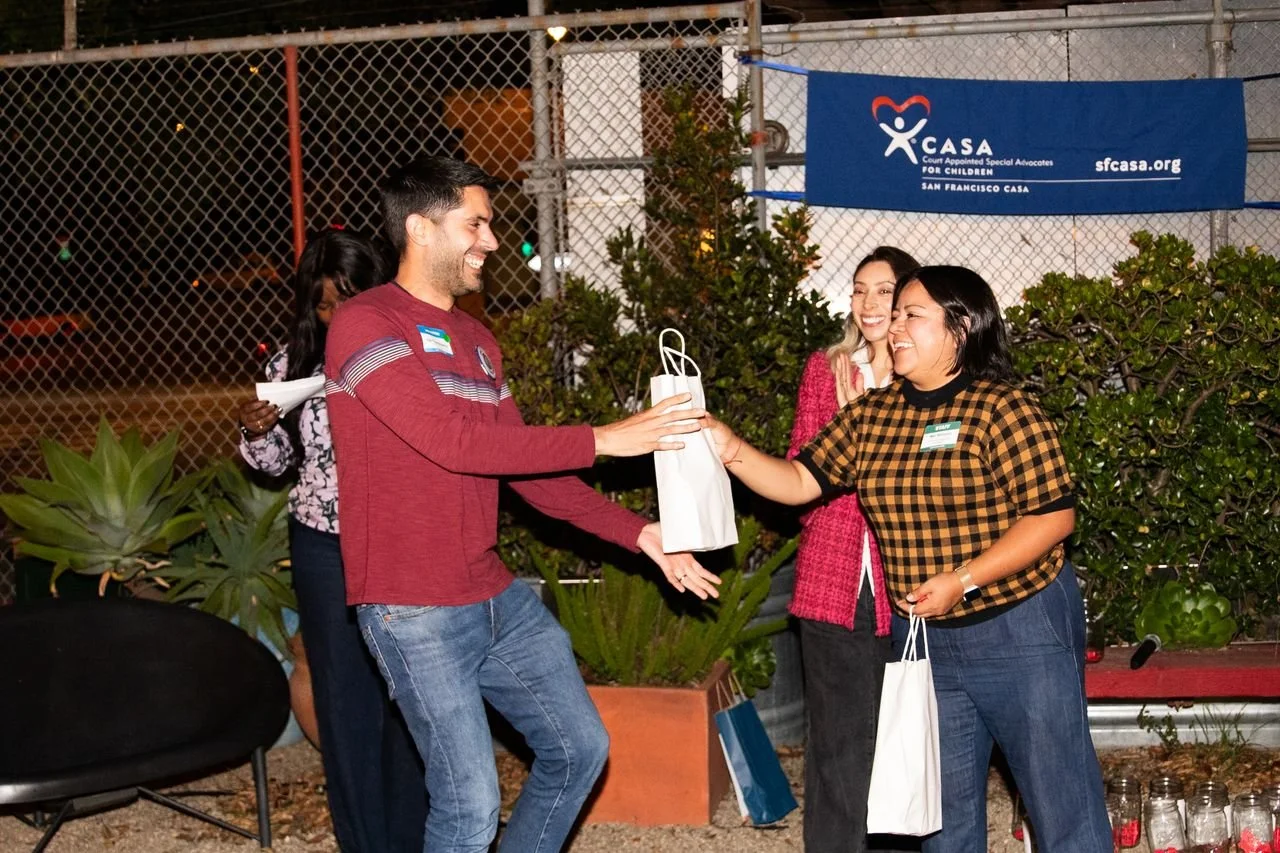 A group of people exchanging a gift bag at an outdoor event, with smiles and friendly interaction, in front of a banner for San Francisco CASA.