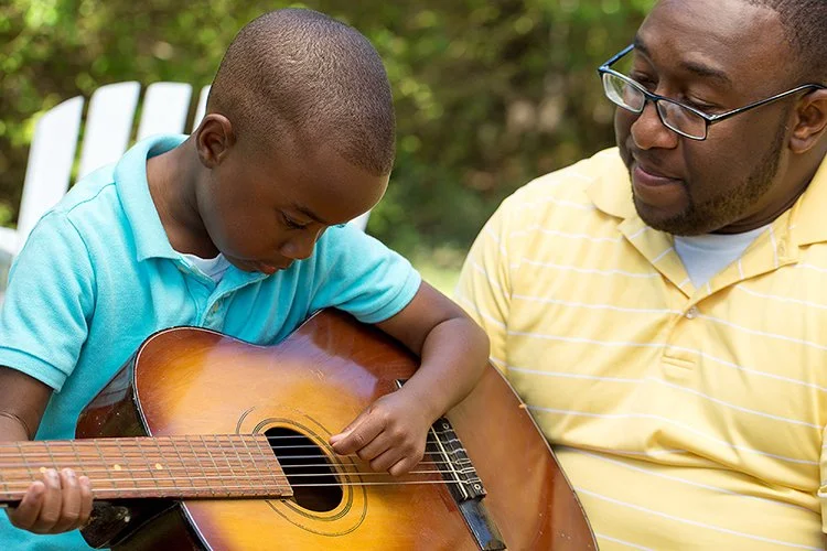 A young boy playing an acoustic guitar while an adult man watches attentively outdoors.