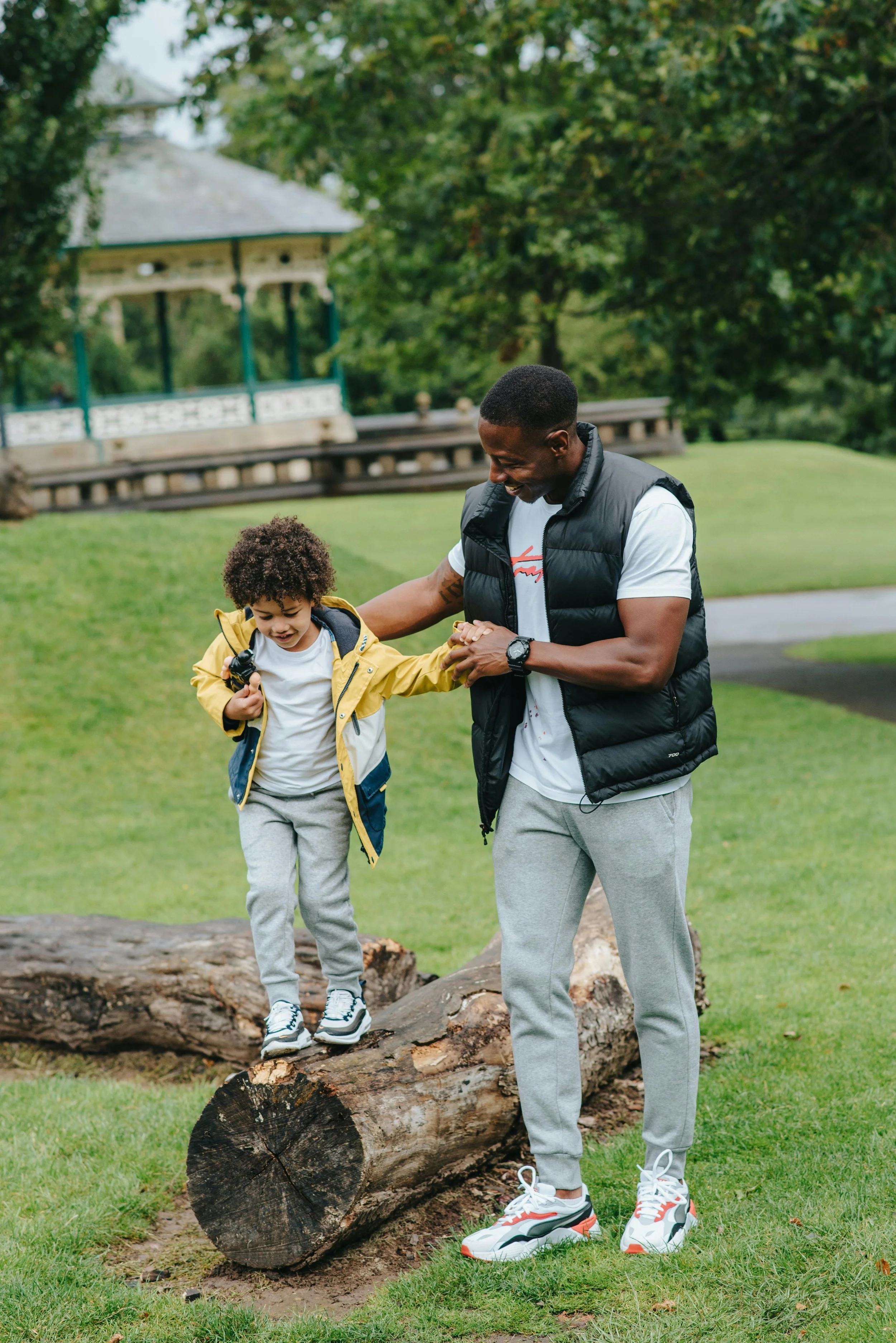 A man helping a young boy walk across a fallen log in a park, with a gazebo and trees in the background.