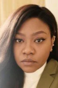 A young woman with long, dark hair, wearing a tan jacket and white blouse, looking into the camera with a neutral expression.
