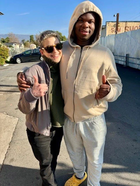 Two people standing outdoors in a parking lot, smiling and giving thumbs up. One is a woman with curly hair wearing sunglasses and a light jacket. The other is a tall man wearing a hooded sweatshirt, sweatpants, and Crocs.