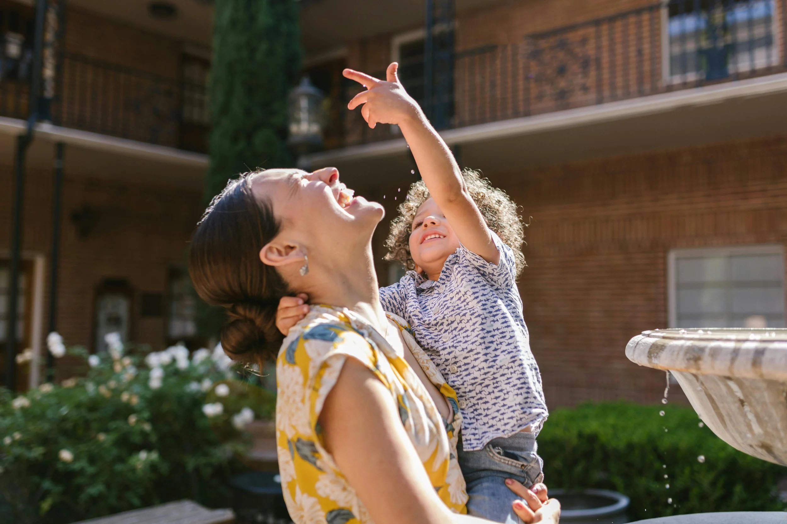 A woman and a young boy enjoying water play outdoors on a sunny day. The woman has dark hair styled in a low bun and is wearing a yellow floral dress. The boy has curly hair and is wearing a patterned shirt. They are smiling and playing near a fountain.