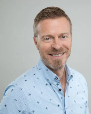 A man with short light brown hair, a beard, and a mustache, smiling, wearing a light blue button-up shirt with small dark patterns, posing against a plain light gray background.