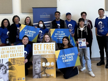 Group of diverse young adults standing in a classroom holding signs that promote free tax help and credit awareness, with some wearing masks.