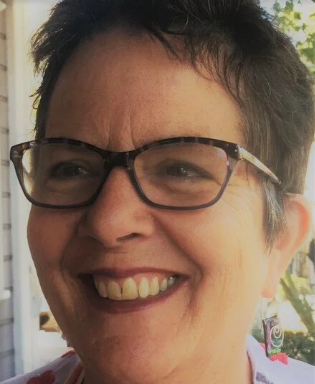 Close-up of a smiling woman wearing glasses, outdoors with greenery in the background.