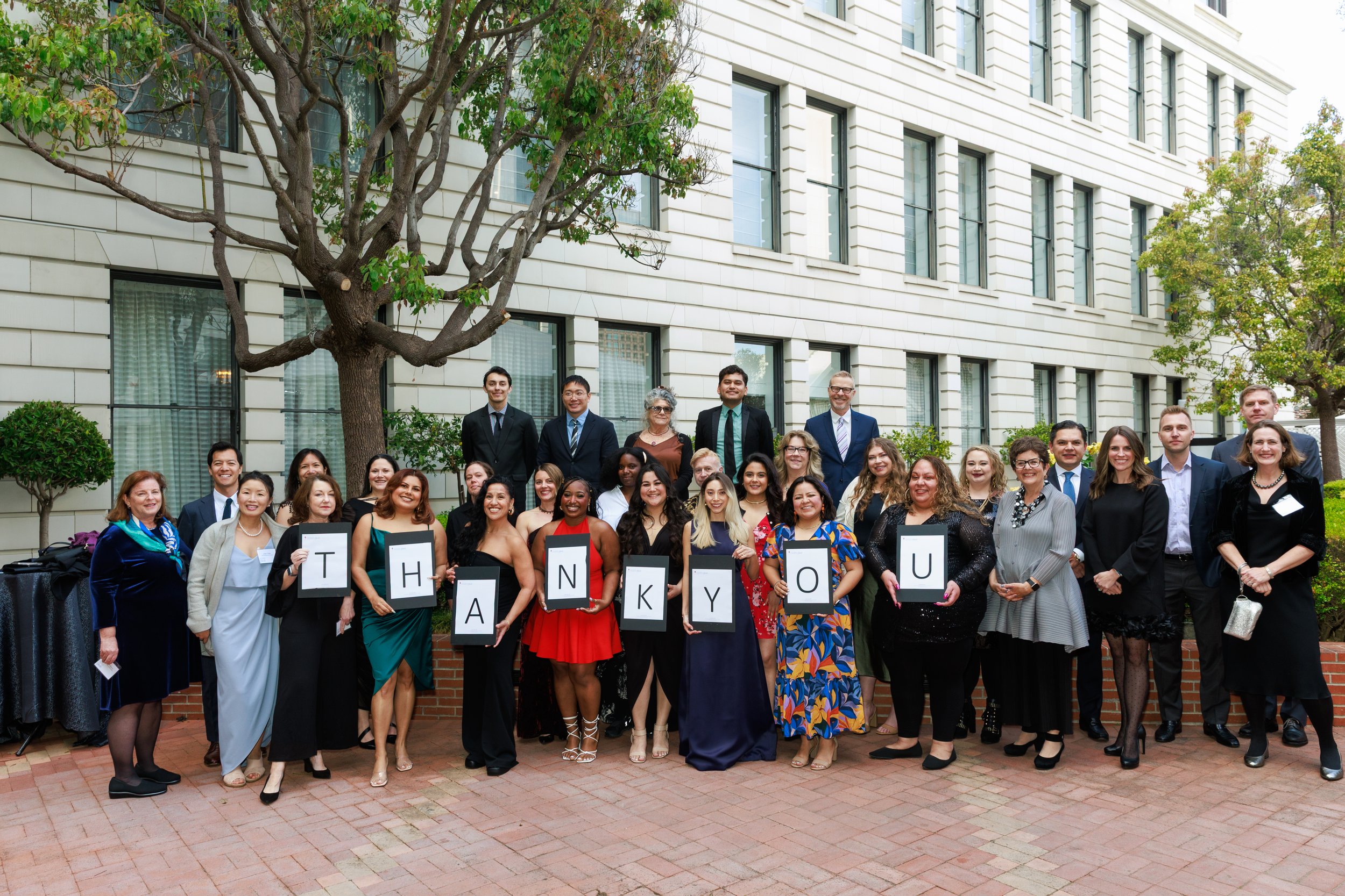 Group of diverse people outdoors celebrating, holding signs spelling 'THANK YOU' in front of a building with trees and windows.