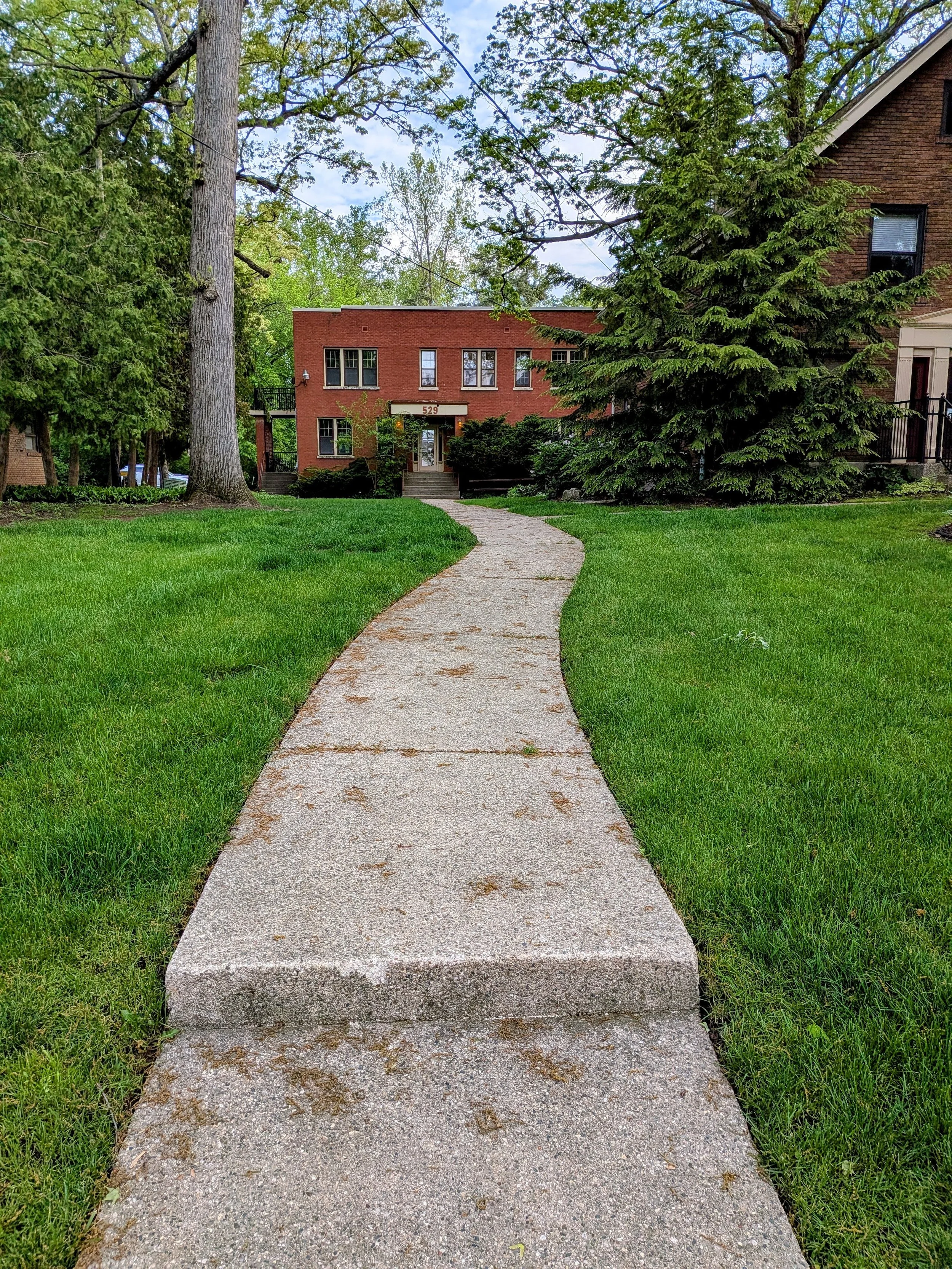 A concrete pathway leading to a brick house with steps at the entrance, surrounded by green grass and trees. Office of Dr. Irene Kraegel, specializing in therapy for anxiety, depression, and chronic pain with Christian Counseling available.
