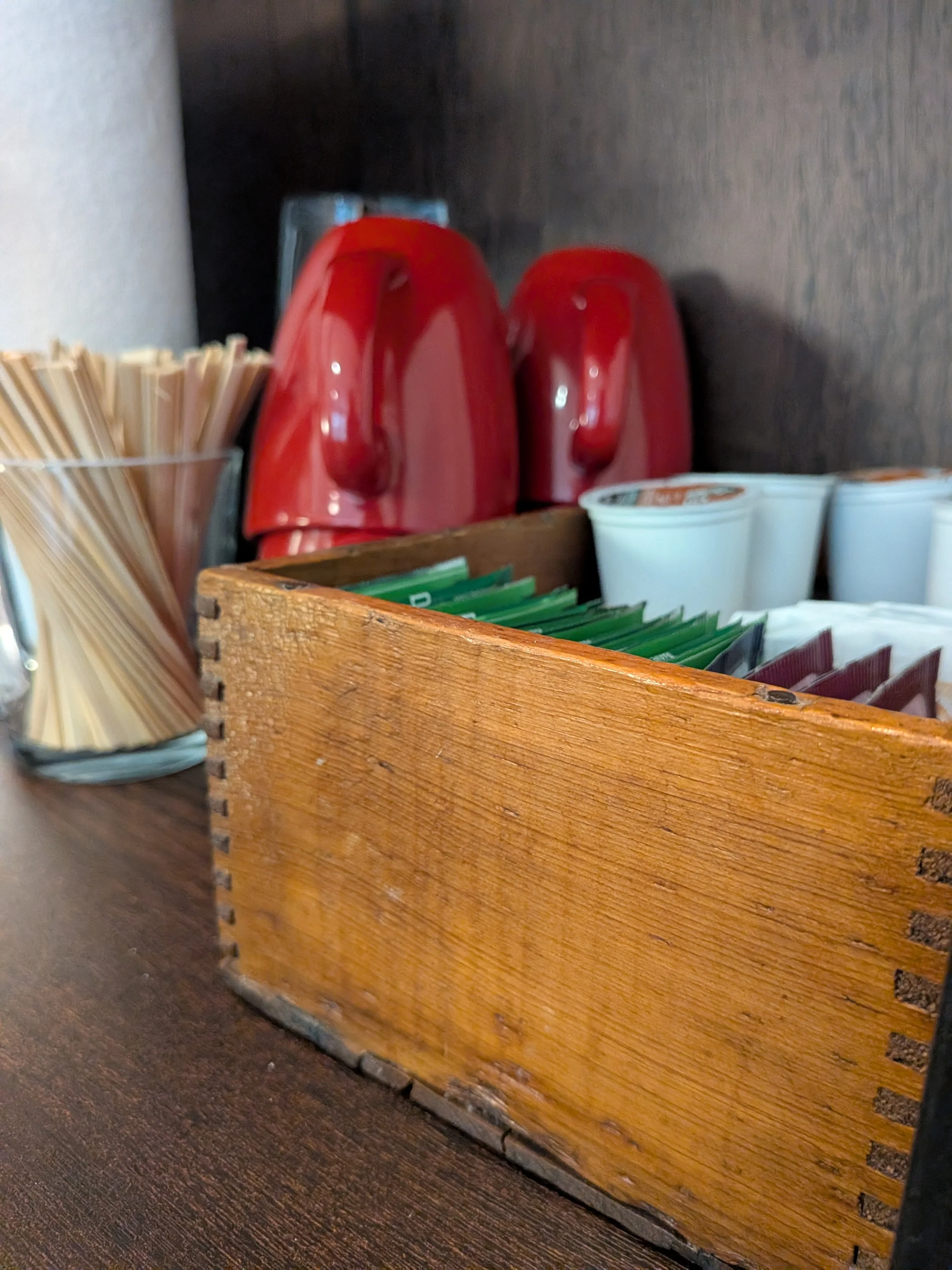 Dark red mugs and glasses are next to a vintage wooden box holding coffee and tea, ready to welcome in-person therapy clients at the office Irene Kraegel in Grand Rapids, MI. Therapy for anxiety, depression, and chronic pain are available.