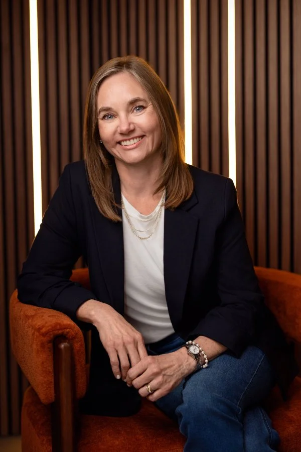 Irene Kraegel, PsyD, smiles with medium-length light brown hair, blue eyes, navy blazer, white tshirt, and gold jewelry, sitting on orange upholstered chair in front of wooden slat wall with lights. Chronic pain Christian therapy in Michigan & online
