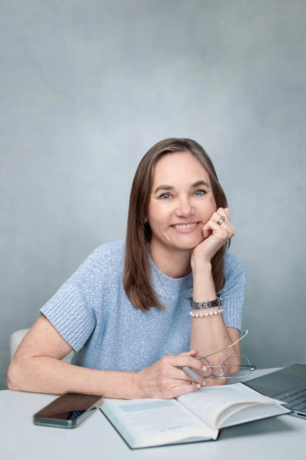 In-person and virtual Christian counseling are available for anxiety/depression and chronic pain with Dr. Irene Kraegel in Grand Rapids, MI. Photo shows Dr. Kraegel smiling and leaning on hand at table with book, phone, computer, and glasses in hand.