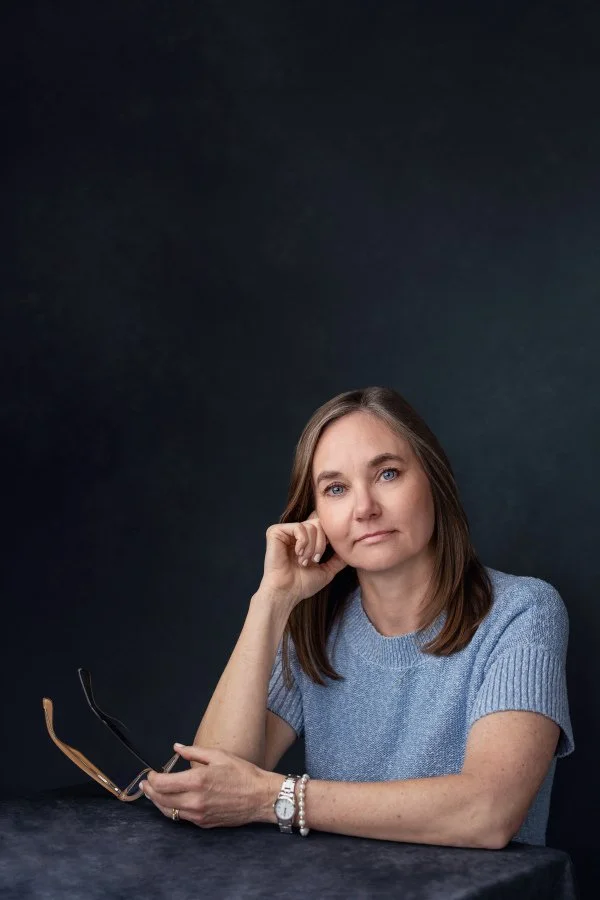 Depression therapy online & in-person Grand Rapids MI from doctoral-level psychologist. Light brown hair & blue eyes sitting at table, resting head on hand, holding glasses in other hand, wearing light blue sweater & looking thoughtfully at camera.