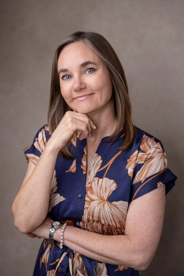 Irene Kraegel provides in-person therapy in Grand Rapids, MI and online in 43 PsyPact states. Shoulder-length brown hair, blue eyes, navy blue dress with large tan & white floral pattern, standing against neutral background.