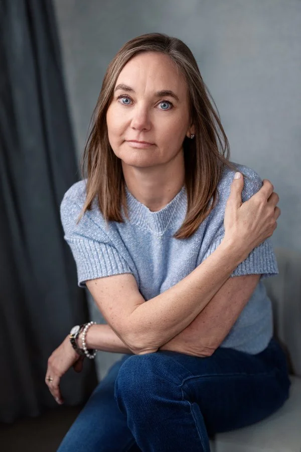 Anxiety therapy in Grand Rapids MI & online with Dr. Irene Kraegel. Shoulder-length brown hair & blue eyes, wearing light blue sweater & dark blue jeans, sitting 1 arm crossed over chest & other resting on knee, looking at camera in softly lit room.