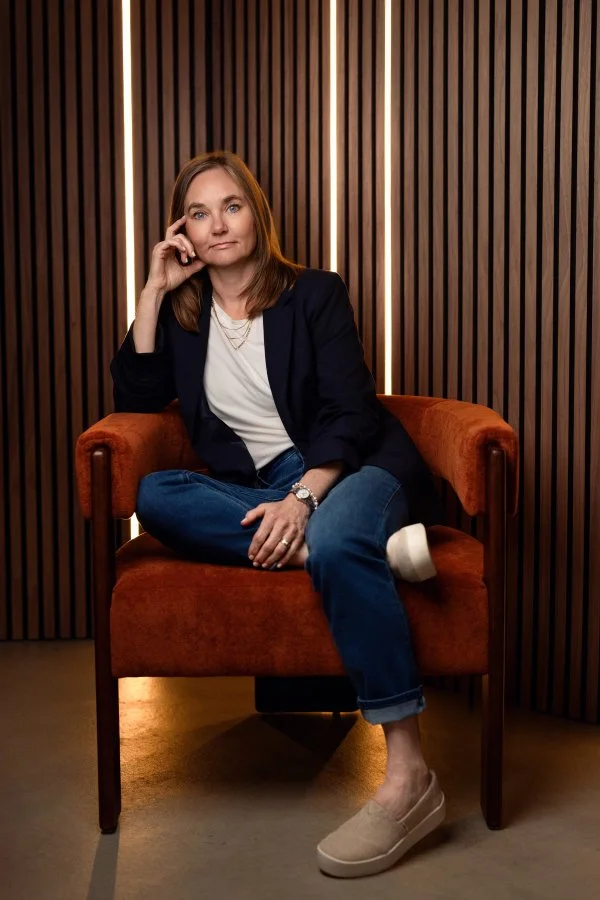 Dr. Irene Kraegel sits in a modern orange chair with wood panel & stripe light background, leaning lightly on fingers with smiling blue eyes, light brown hair, & one leg pulled up under the other. Christian counseling in-person in Michigan and online