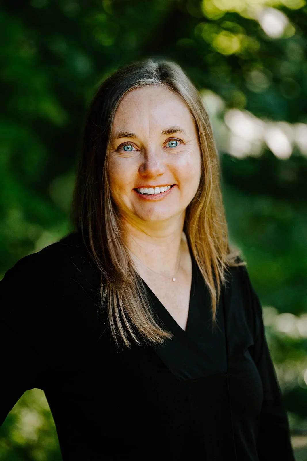 Irene Kraegel, PsyD stands in front of green trees wearing a black v-neck shirt with dark brown hair, blue eyes, and a smile. Dwell Psychological Services provides Christian counseling in-person in Grand Rapids, MI and online in 43 PsyPact states.