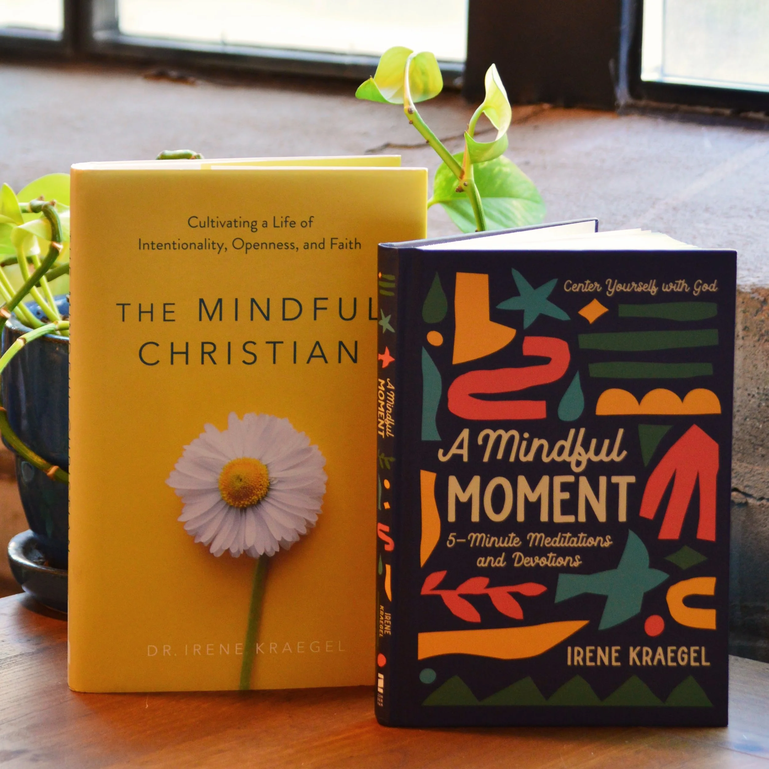 Two books on a wooden surface near a window, with a green plant nearby. The book on the left has a yellow cover with a daisy flower and is titled 'The Mindful Christian' by Dr. Irene Kraegel. The book on the right has a dark, colorful cover titled 'A Mindful Moment: 5-Minute Meditations and Devotions' by Irene Kraegel.