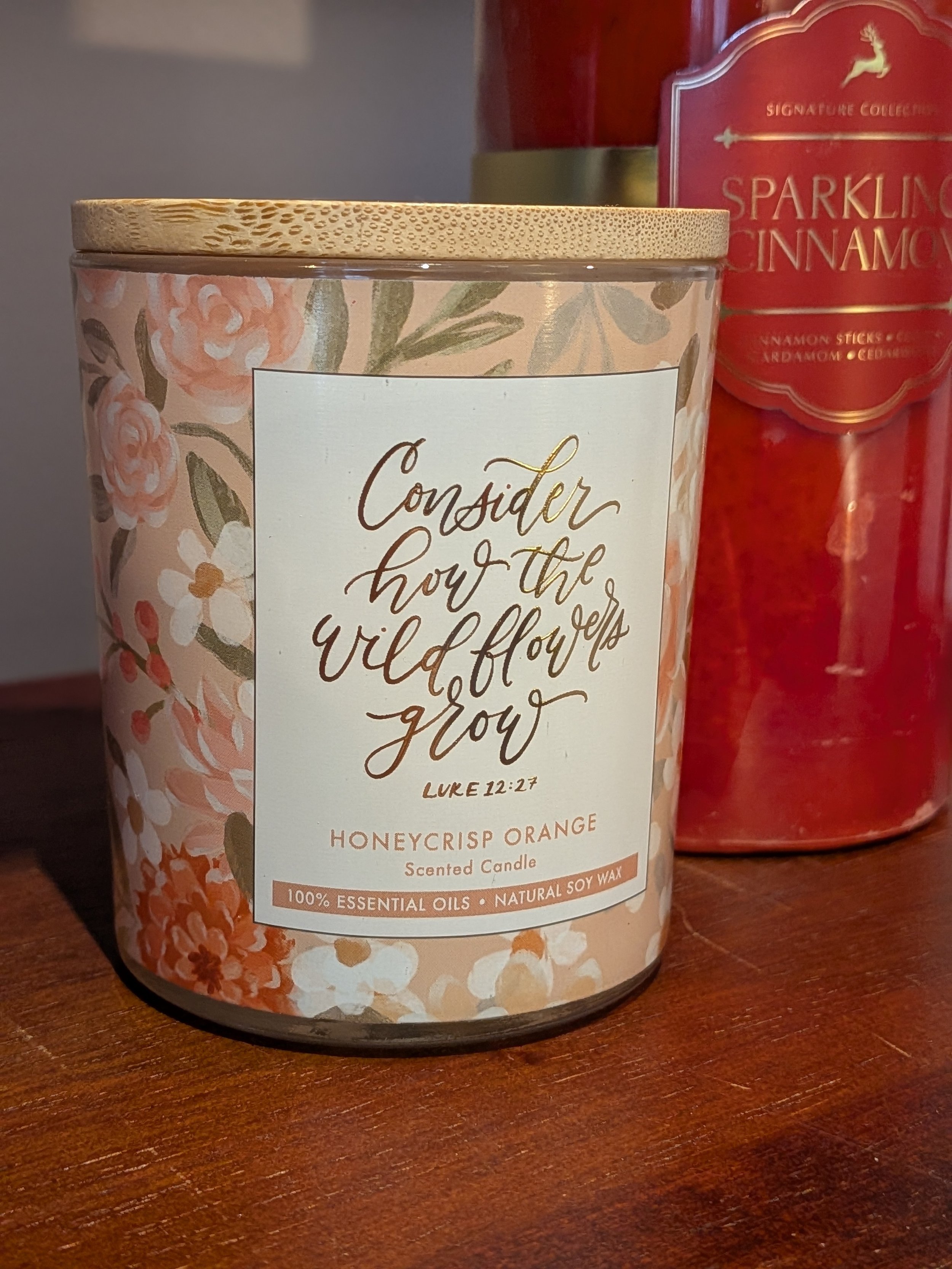 A scented candle in therapy office of Dr. Irene Kraegel, PsyD in Grand Rapids, MI with a floral design and a label that reads 'Consider how the wildflowers grow' with scripture Luke 12:27, in a pink and orange floral pattern, next to a red candle con