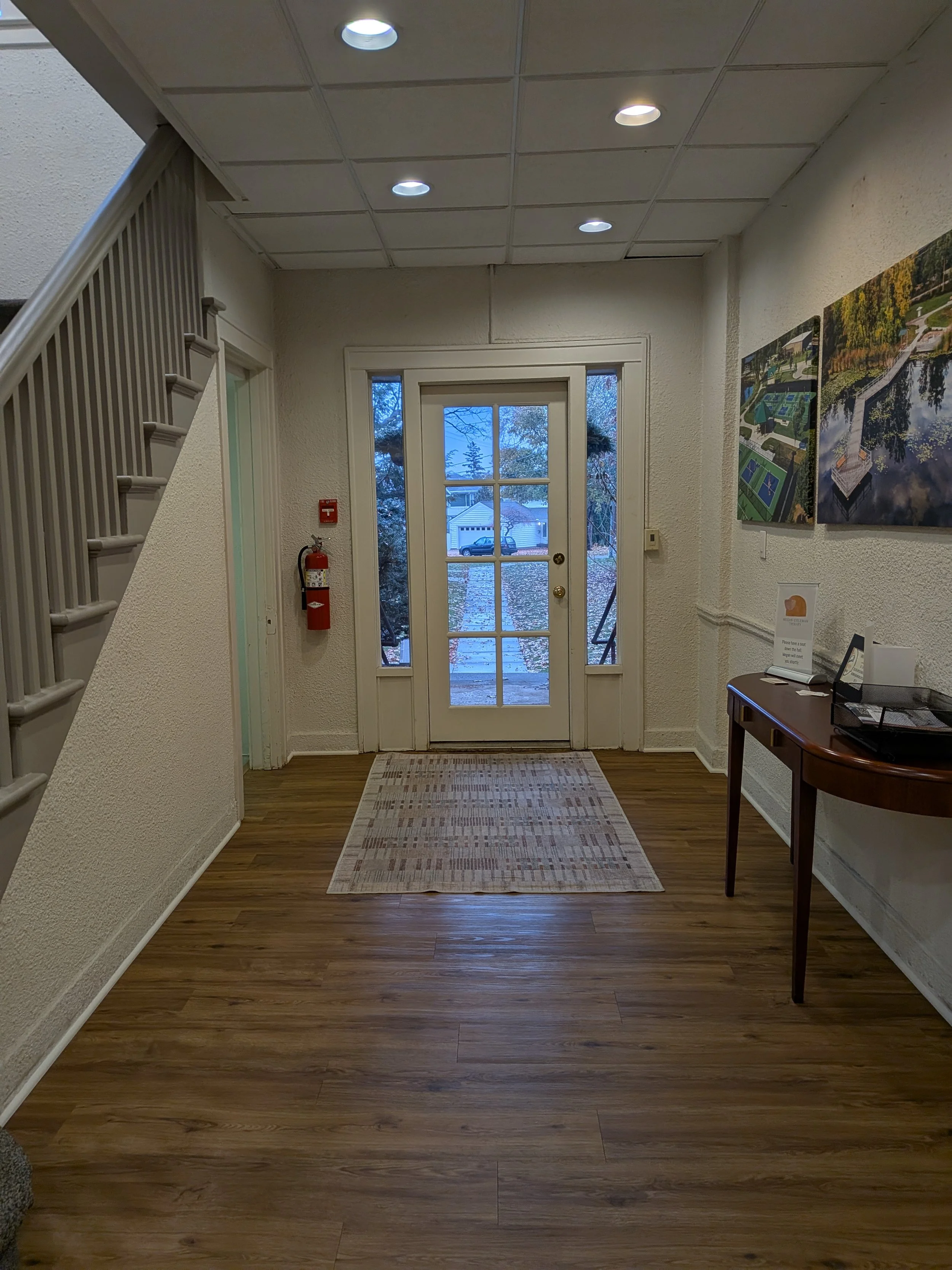 Therapy office entryway of Dr. Irene Kraegel, PsyD, in East Grand Rapids, MI with a glass-paneled door, wooden flooring, a small rug, wall art, a side table with a tray, a fire alarm, and a staircase on the left.