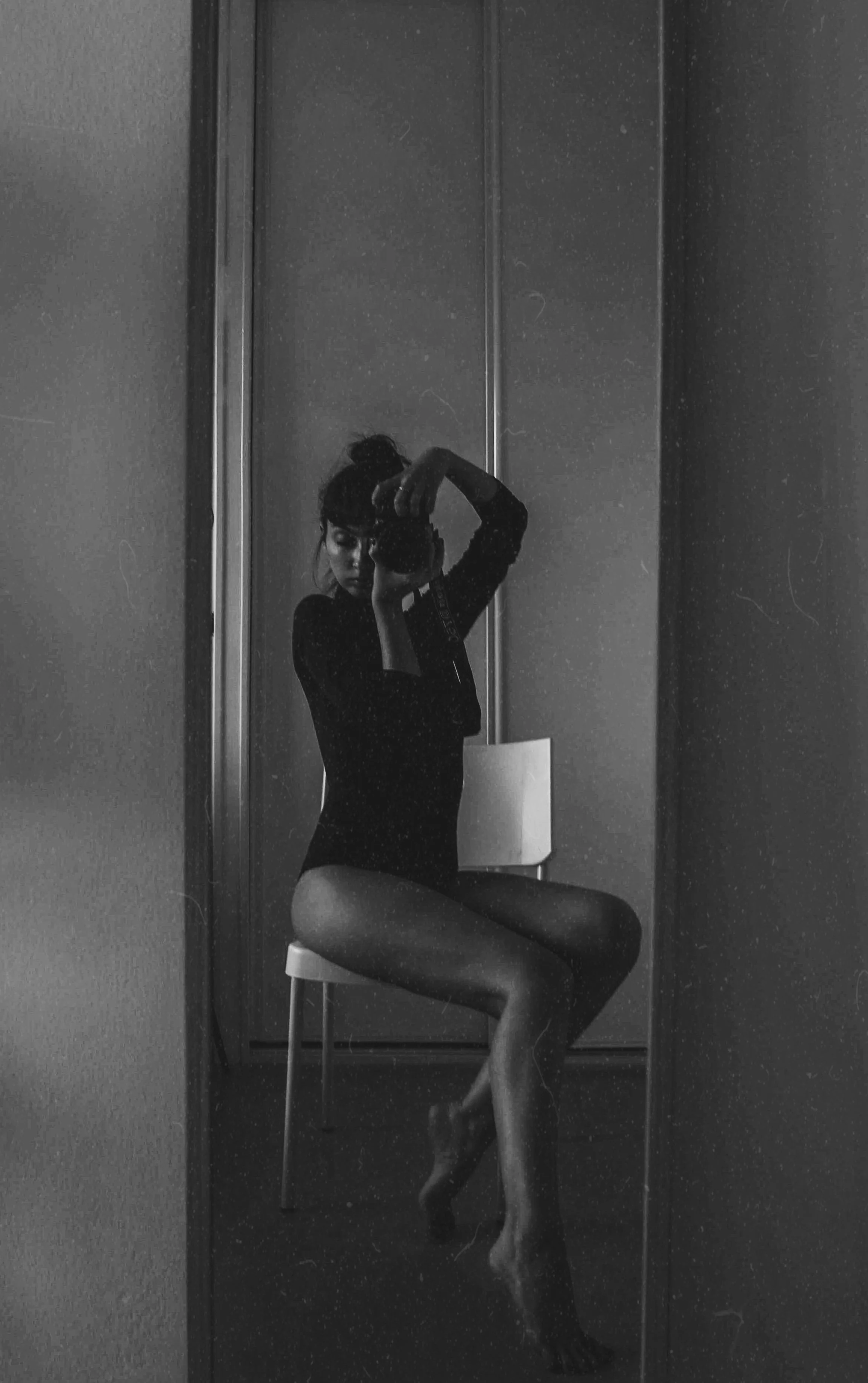 A woman taking a selfie in front of a mirror, sitting on a chair with her legs crossed, in black and white.