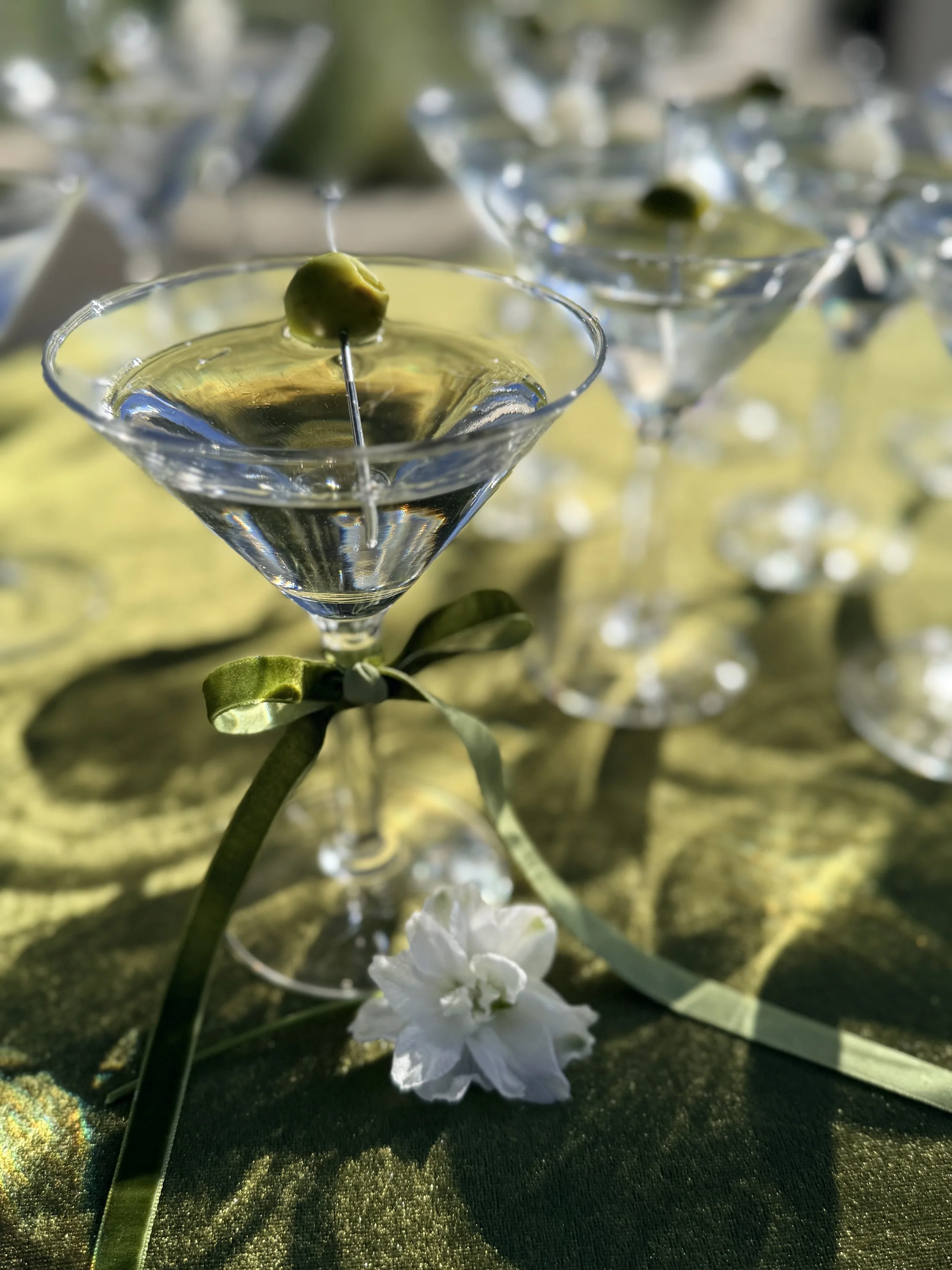 Close-up of a martini glass filled with a clear liquid, garnished with a green olive on a skewer, set on a green fabric surface with white flowers and blurred similar glasses in the background.