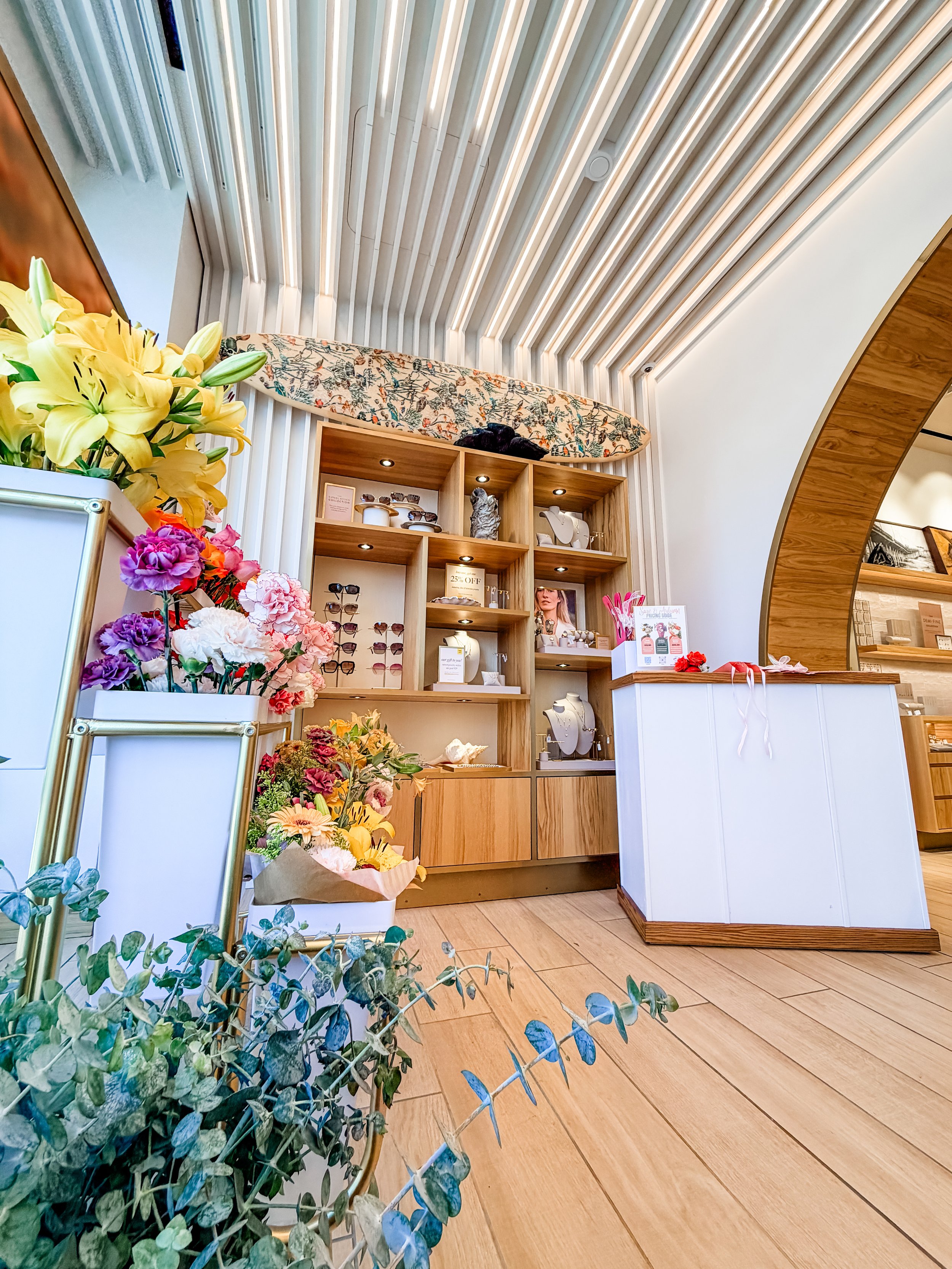 Interior view of a jewelry or accessory store with wooden display shelves, floral arrangements, and a hanging surfboard with a colorful pattern on the wall behind the shelves, and a curved wooden ceiling.