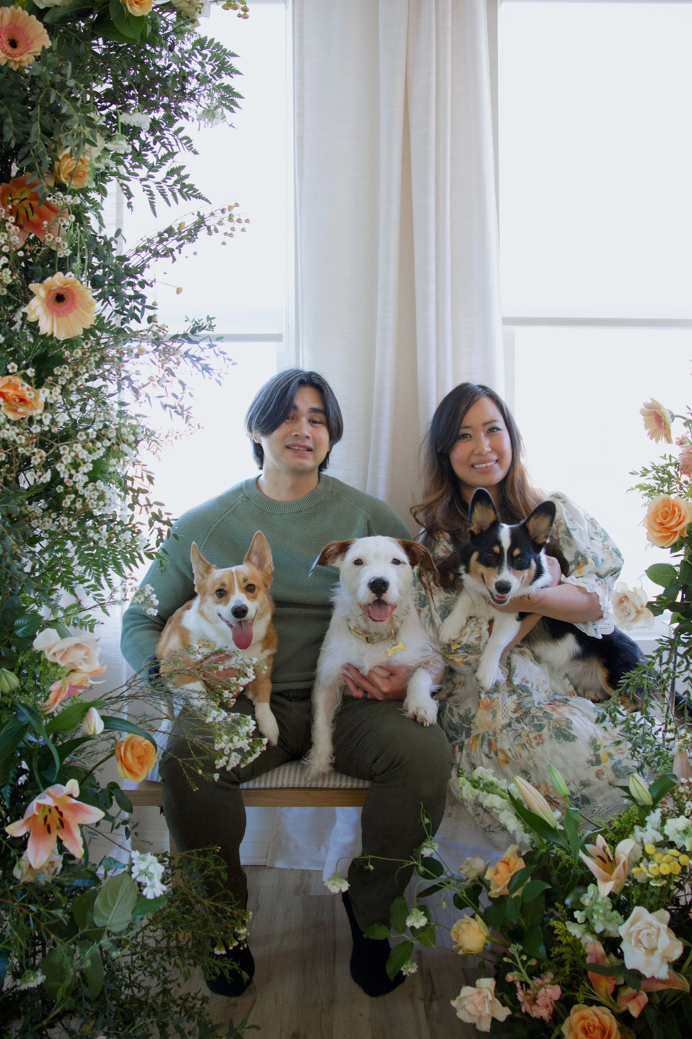 A smiling couple sitting on a bench with three dogs surrounded by an arrangement of colorful flowers in front of a bright window. San Diego Wedding Florals