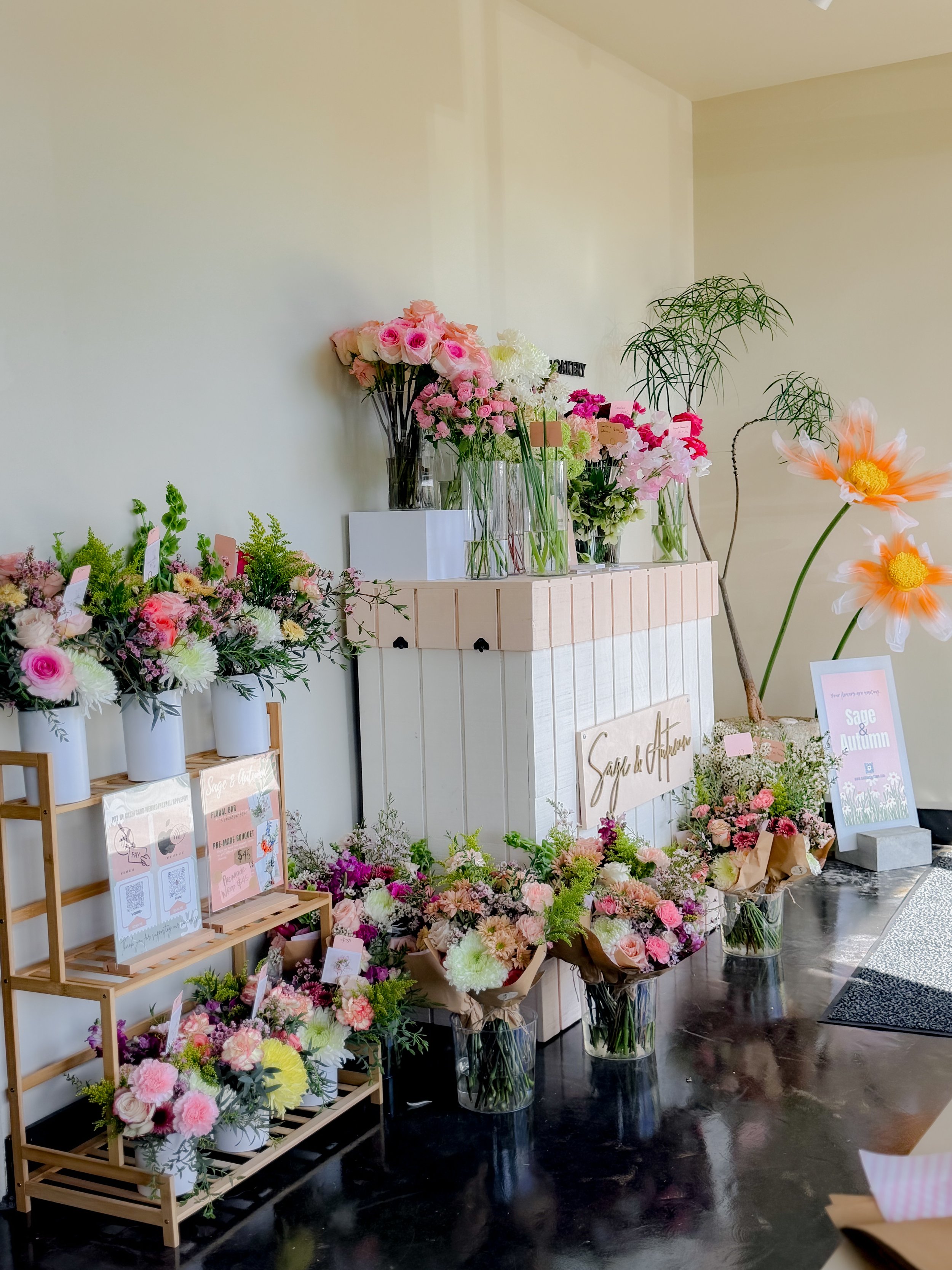 A display of various colorful fresh flowers in glass vases and a wicker basket on white wooden furniture, with a large floral arrangement on a white and beige wooden box. Floral Bar San Diego 