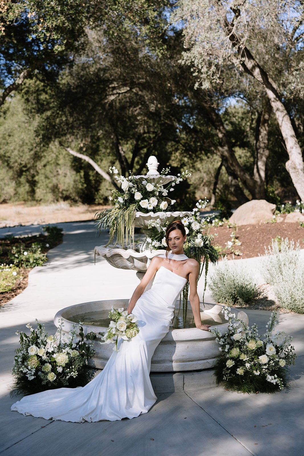 A woman in a white strapless wedding dress and matching neck accessory sitting on a fountain decorated with white flowers, with more white floral arrangements around the base, outdoors under trees.