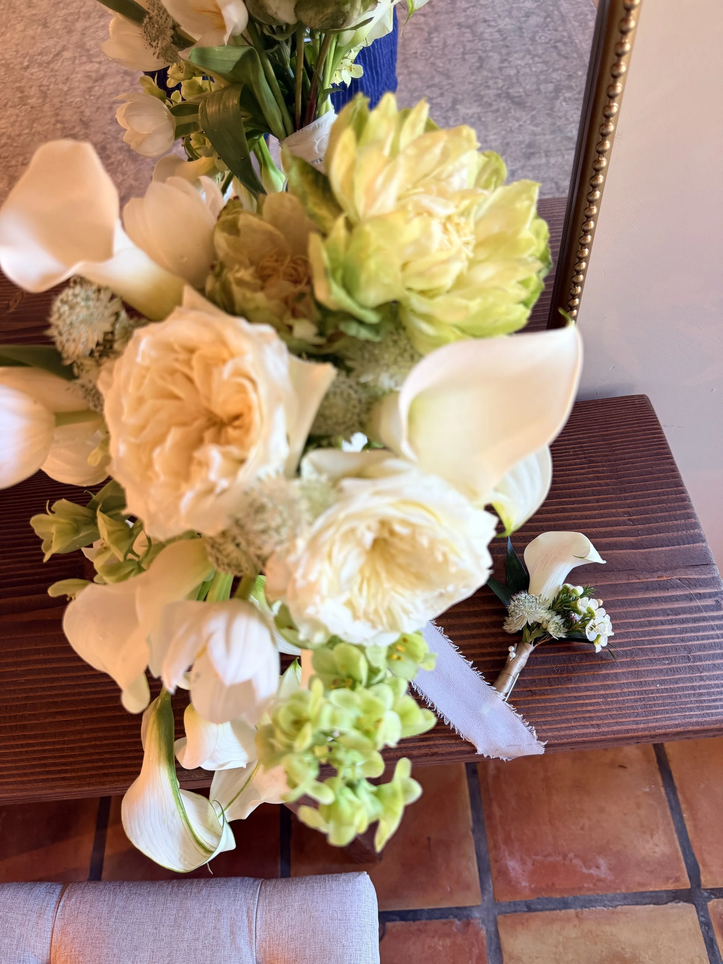 A bouquet of white and pale yellow flowers, including calla lilies, roses, and other blooms, arranged on a wooden surface with a mirror and a boutonniere nearby.