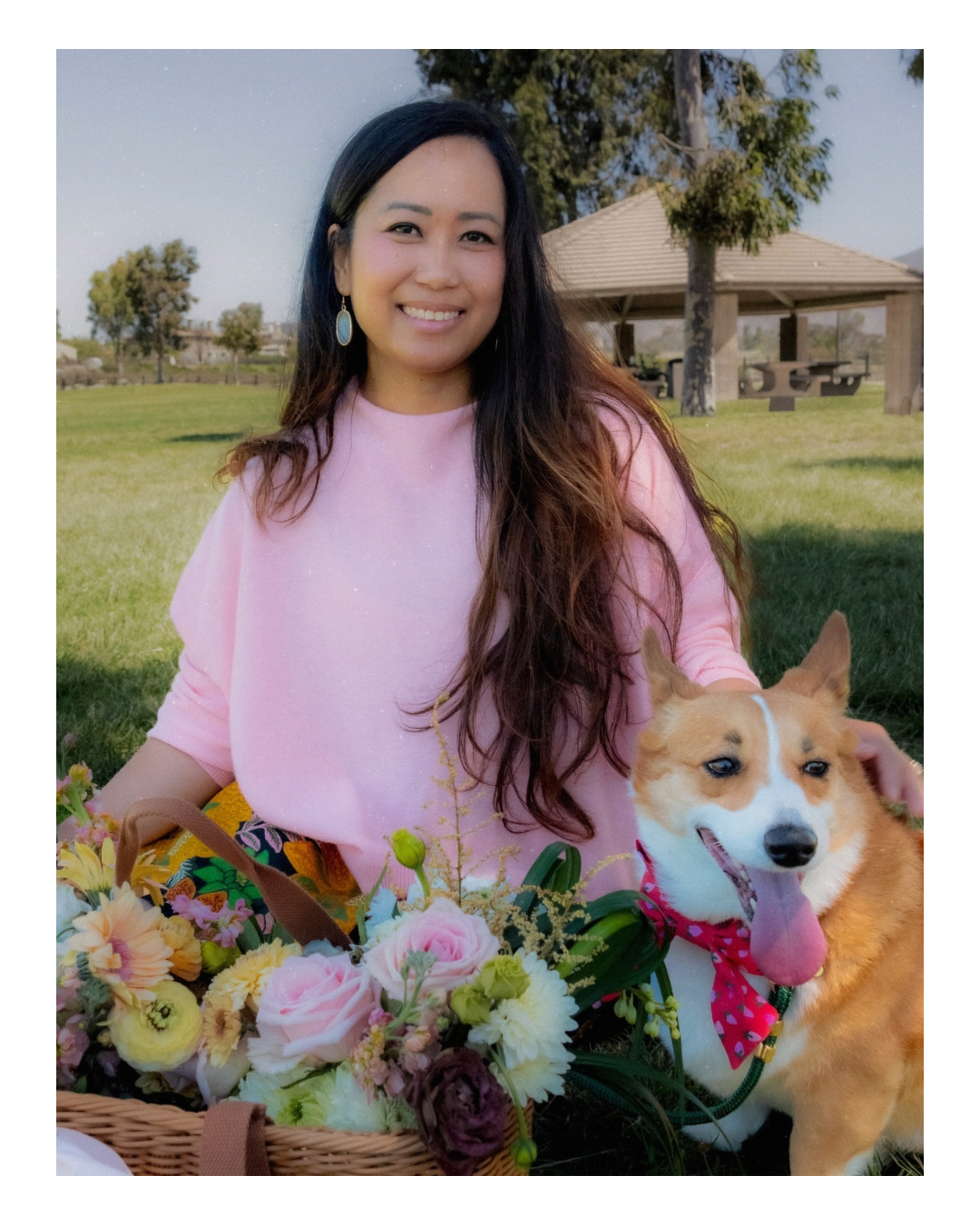 A woman with long dark hair, wearing a pink top and blue earrings, sitting on a grassy park with a smiling dog wearing a red bandana, surrounded by a basket of colorful flowers, with a gazebo and trees in the background.