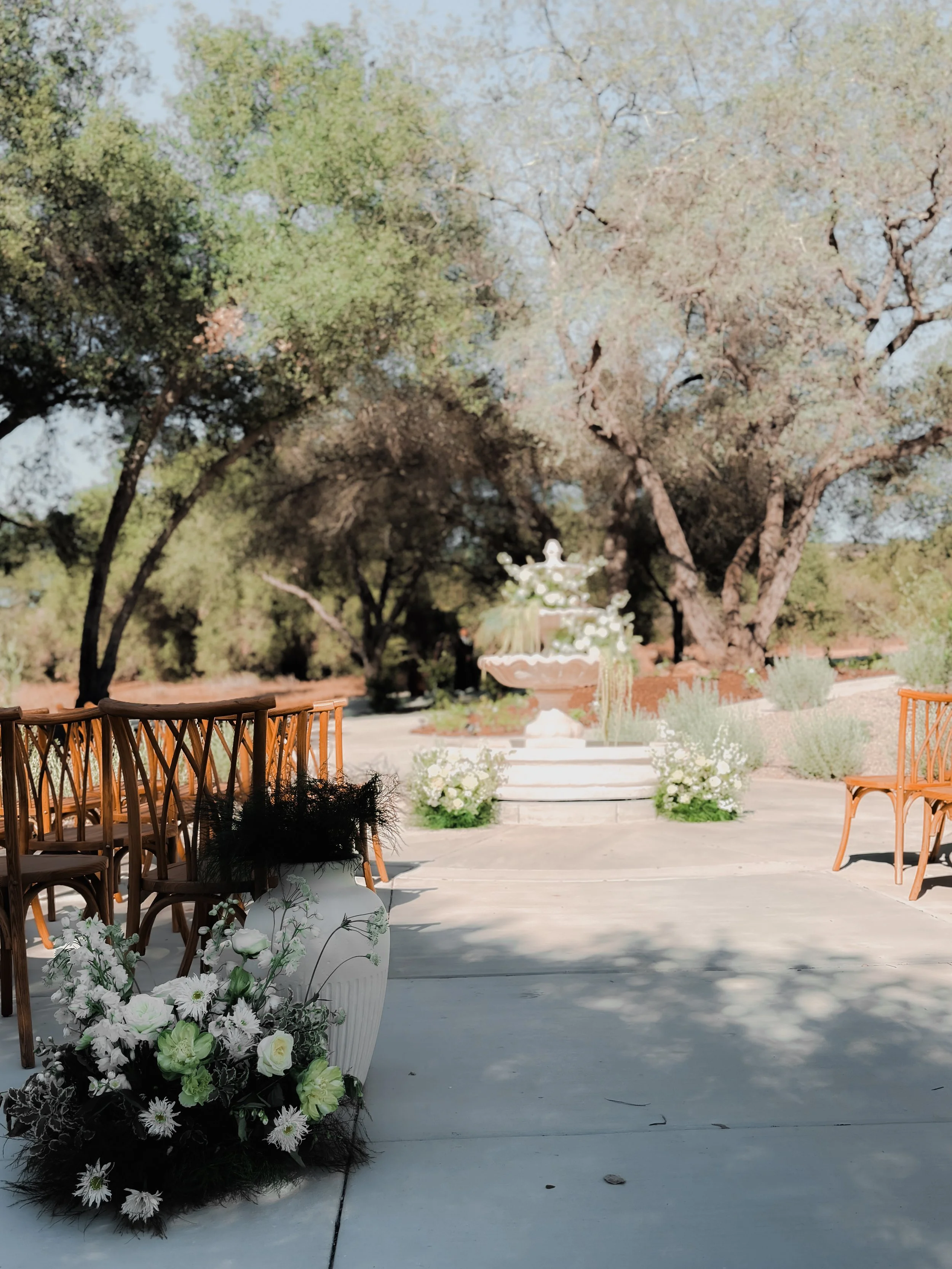 Outdoor wedding ceremony setup with wooden chairs, floral arrangements in vases, a stone fountain, and large trees in the background.