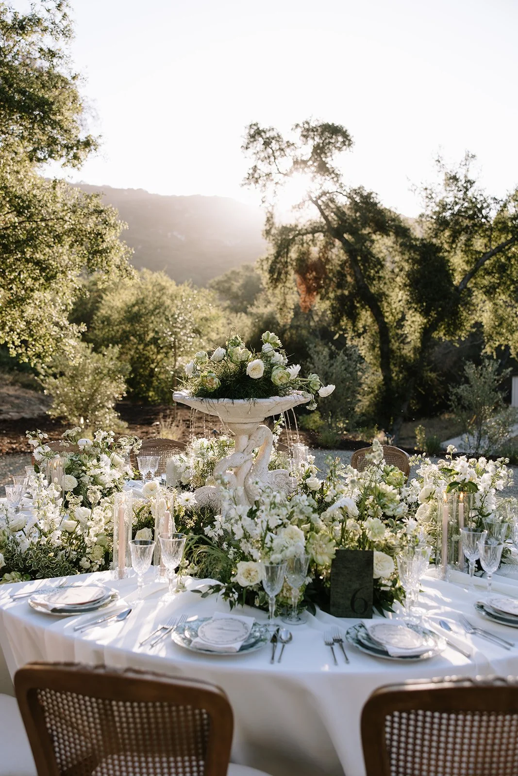 A beautifully decorated outdoor event table with a white tablecloth, floral arrangement, and dinnerware, set in a garden during sunset.