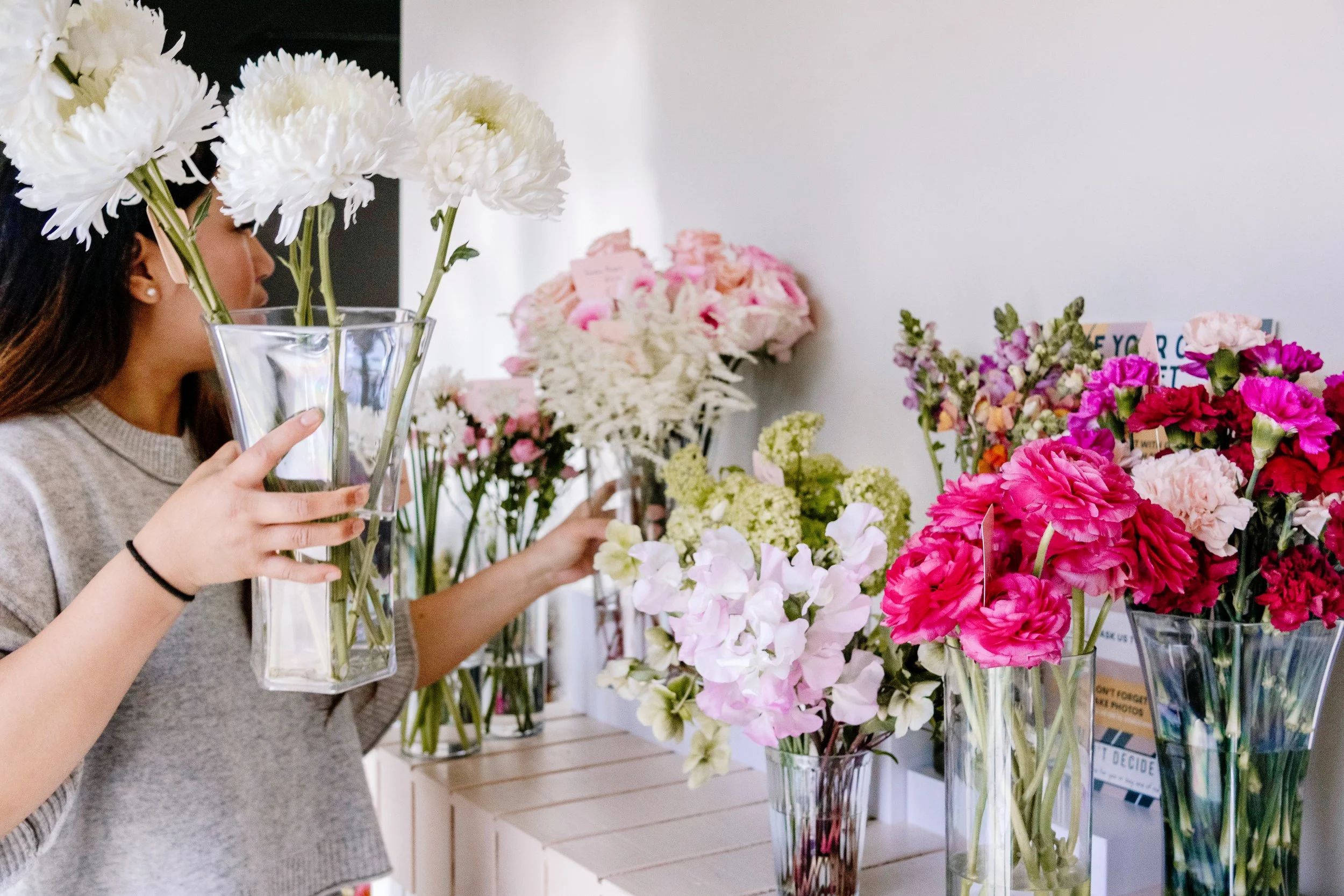 A flower-themed outdoor setup with artificial flowers, a white and peach-striped stand, a small white and peach-striped table with a "Sage & Autumn" sign, large daisies with orange centers, and a backdrop of a fence, blue sky, and clouds.