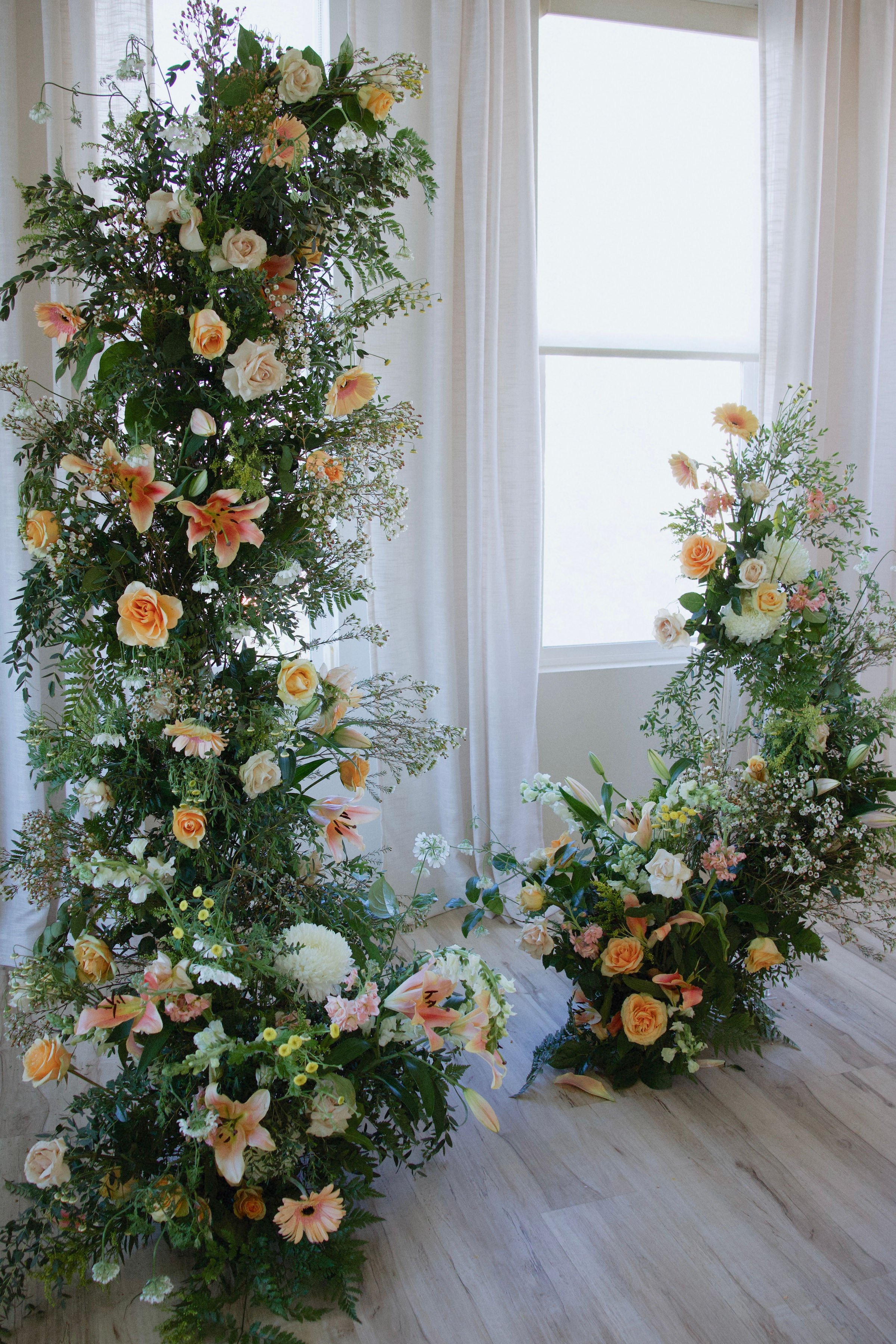 Two floral arrangements with various light-colored flowers, greenery, and some pink accents, set against a backdrop of white curtains and a window.