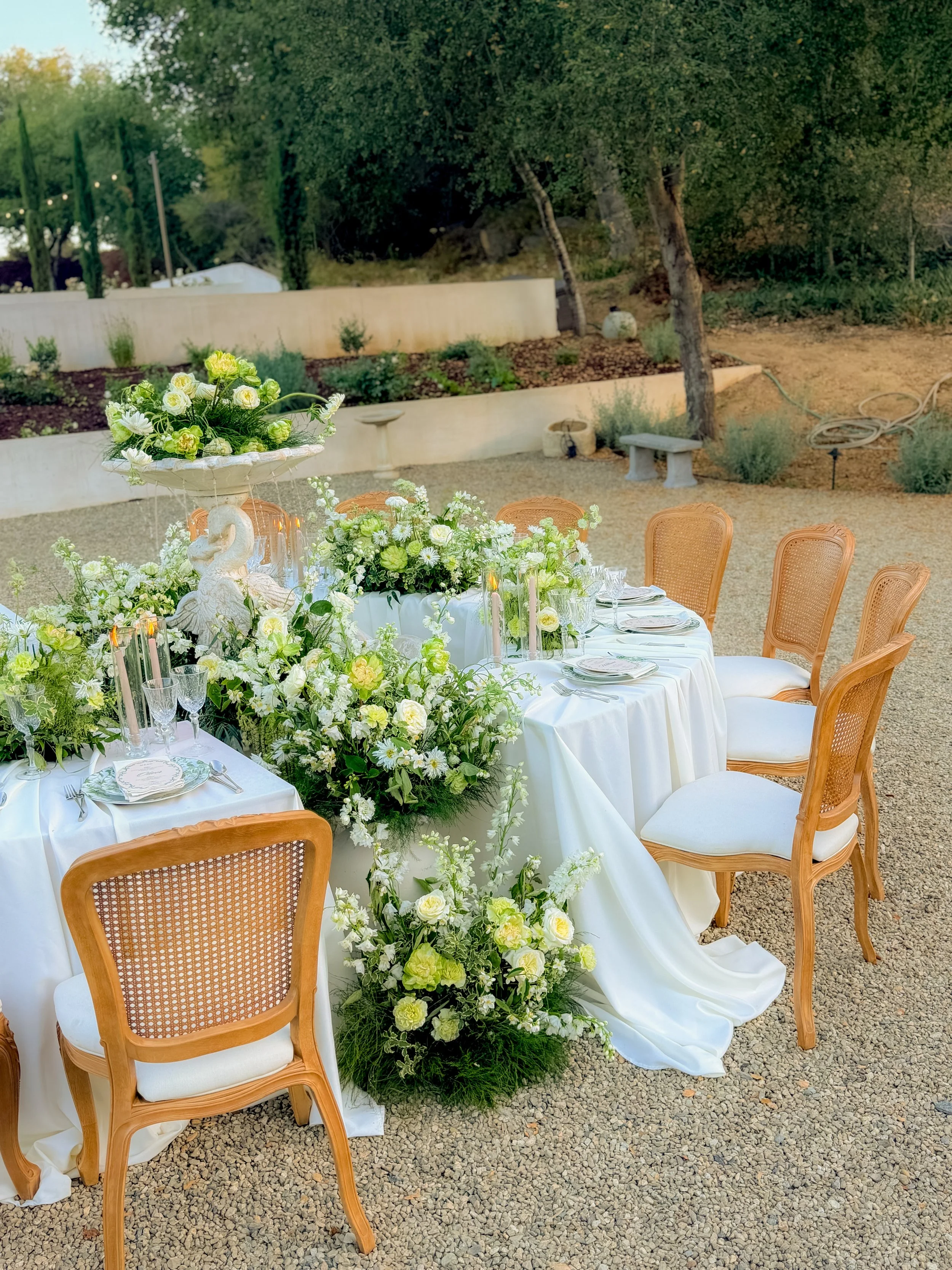 Elegant outdoor dining table decorated with white and green floral arrangements, set with plates, glasses, and candles, surrounded by wooden chairs on a gravel surface with trees in the background.