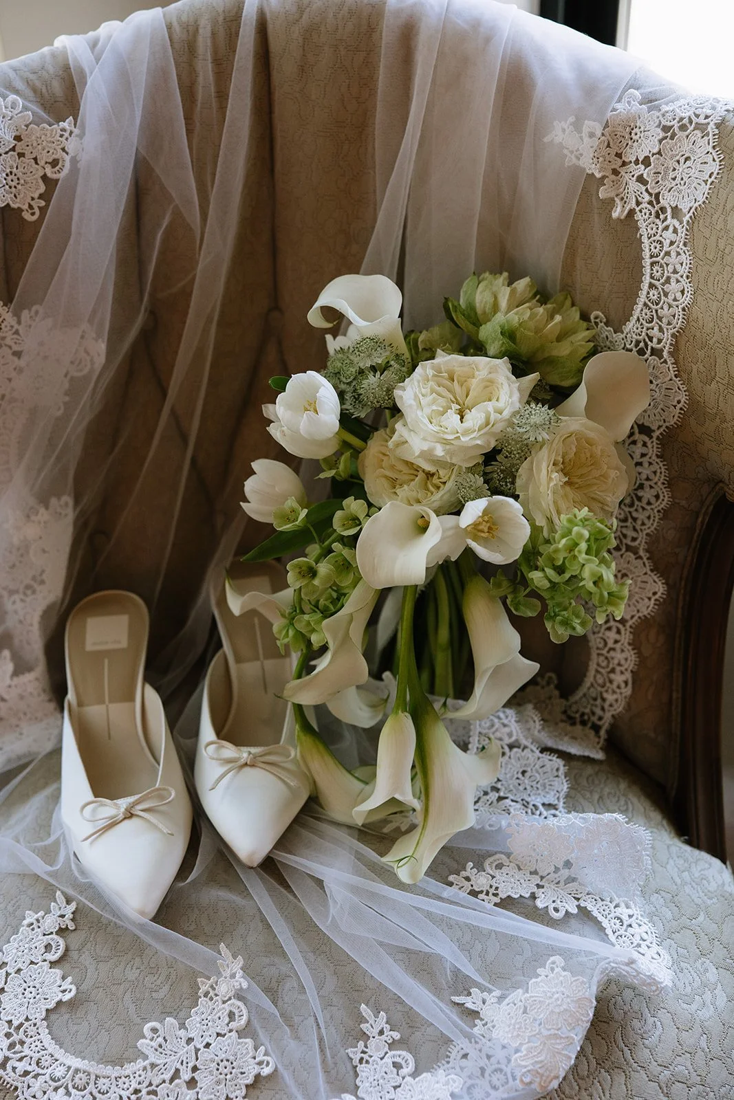 A bridal bouquet of white flowers, including calla lilies, roses, and ranunculus, resting on lace fabric with a pair of white satin shoes nearby, decorated with lace and ribbons.