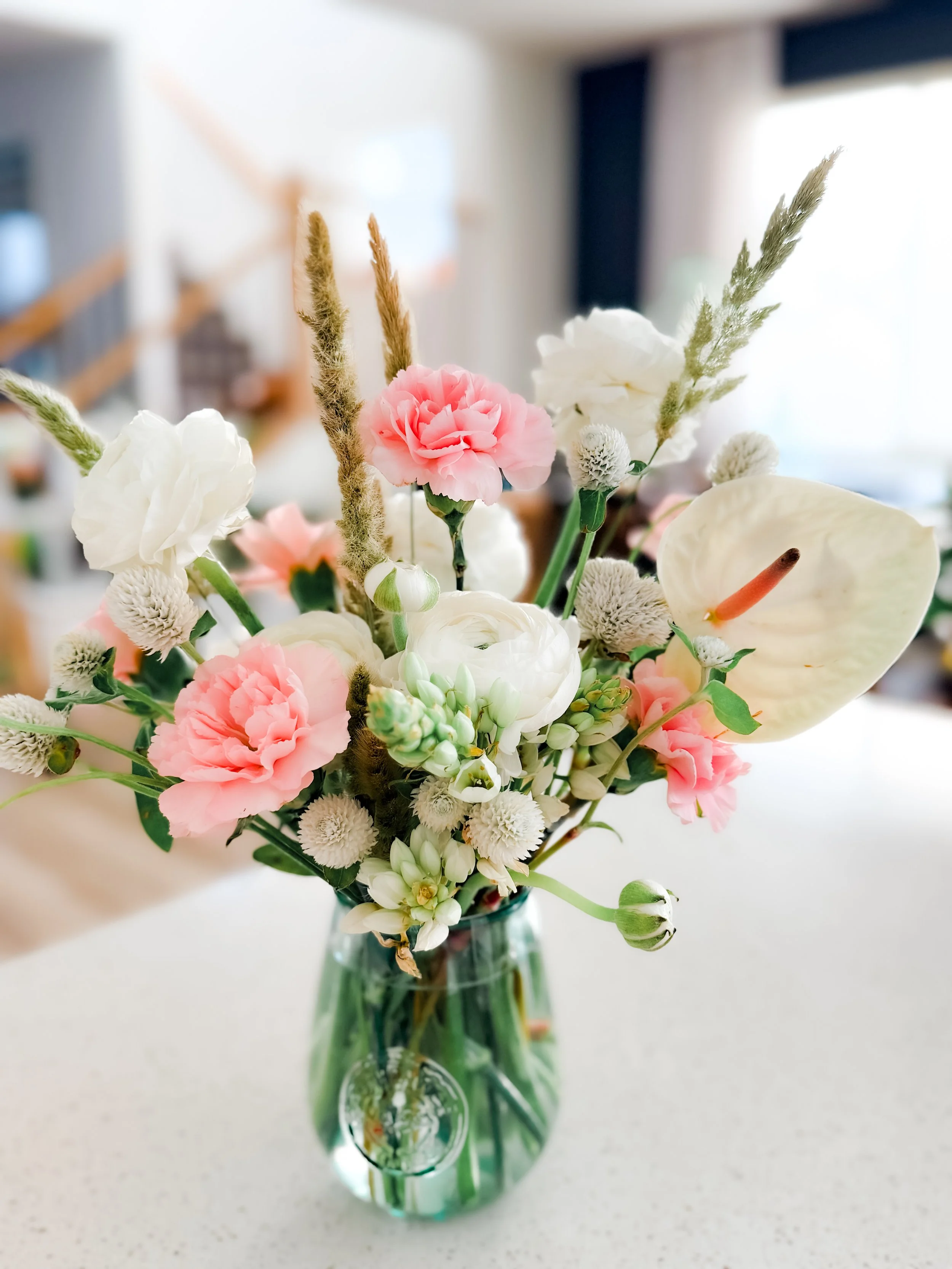 A bouquet of mixed flowers in a green glass vase on a white surface, with a blurred background.