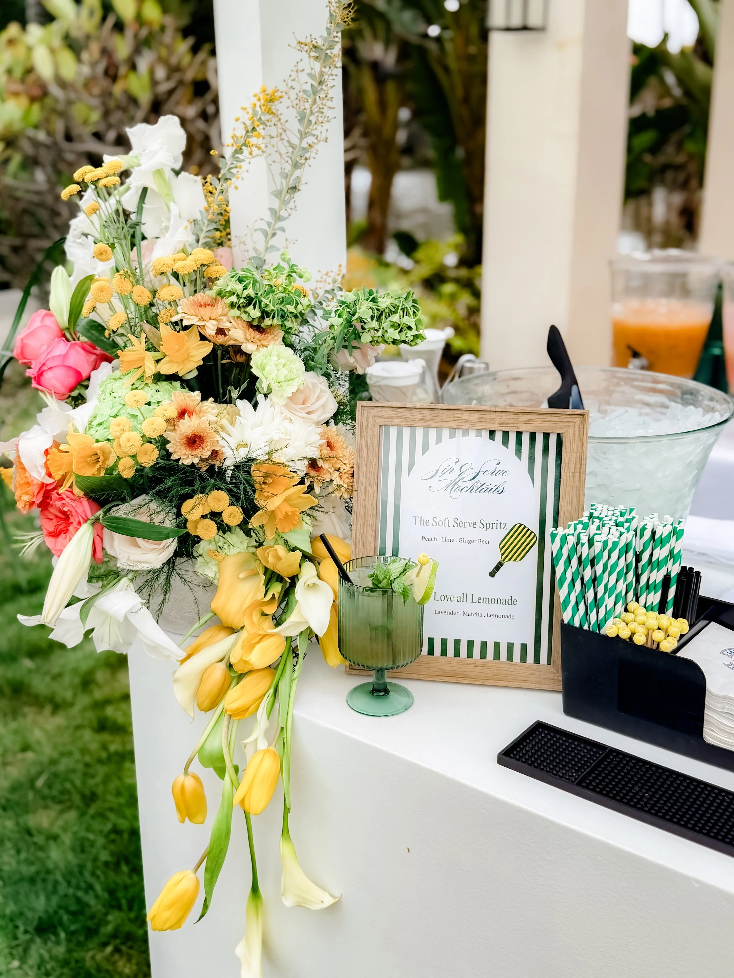 A floral arrangement with various colorful flowers, a framed menu for cocktails and lemonade, striped straws, and plastic cups with drinks on a white table at an outdoor event.