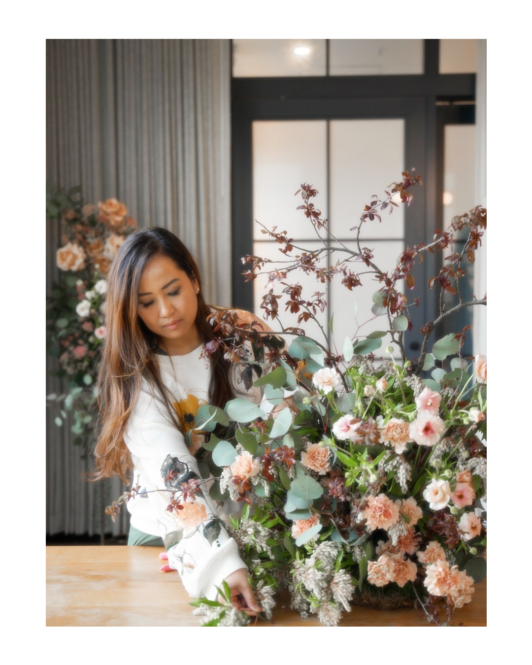 Woman arranging a large floral arrangement with pink, peach, and white flowers and green leaves in a room with light-colored curtains and a window.