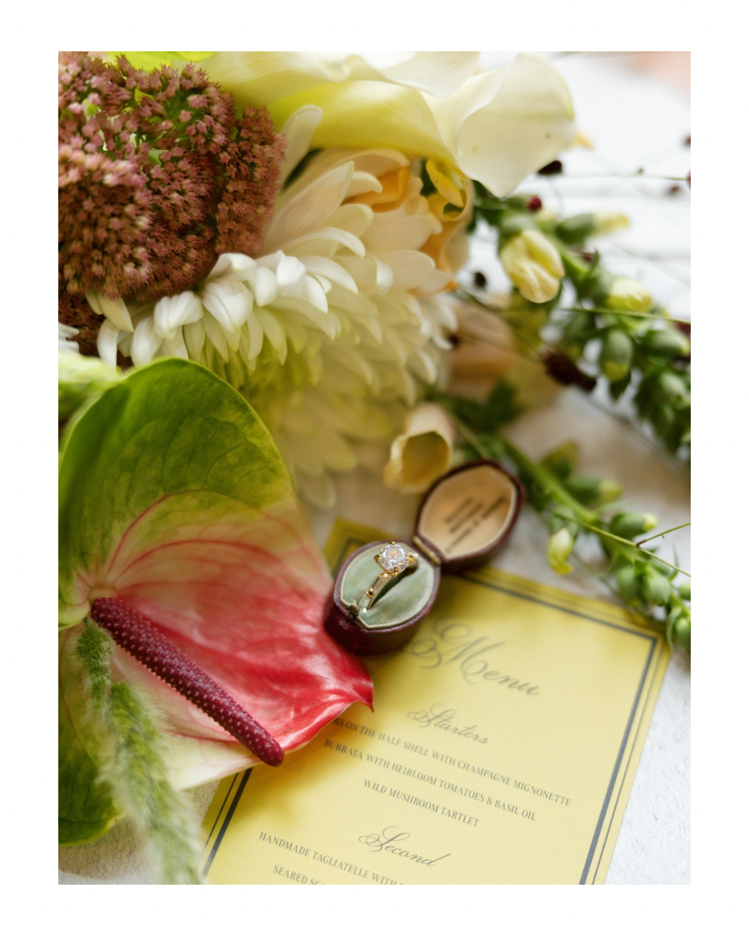 Close-up of a wedding bouquet with white, pink, and green flowers, a diamond engagement ring in a velvet box, and a yellow menu card.