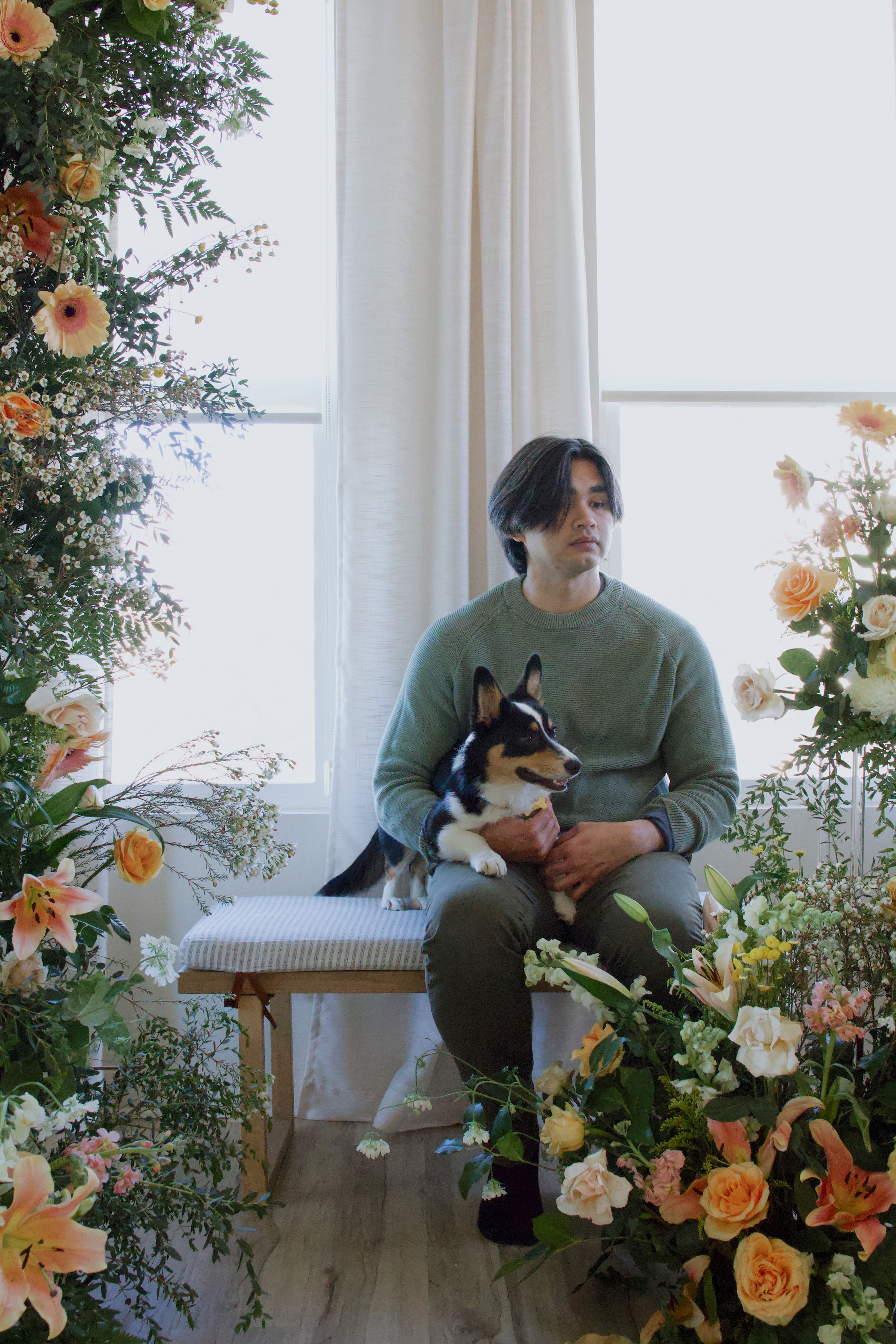 A man sitting on a bench near a window, holding a black and white dog, surrounded by flowers and greenery.