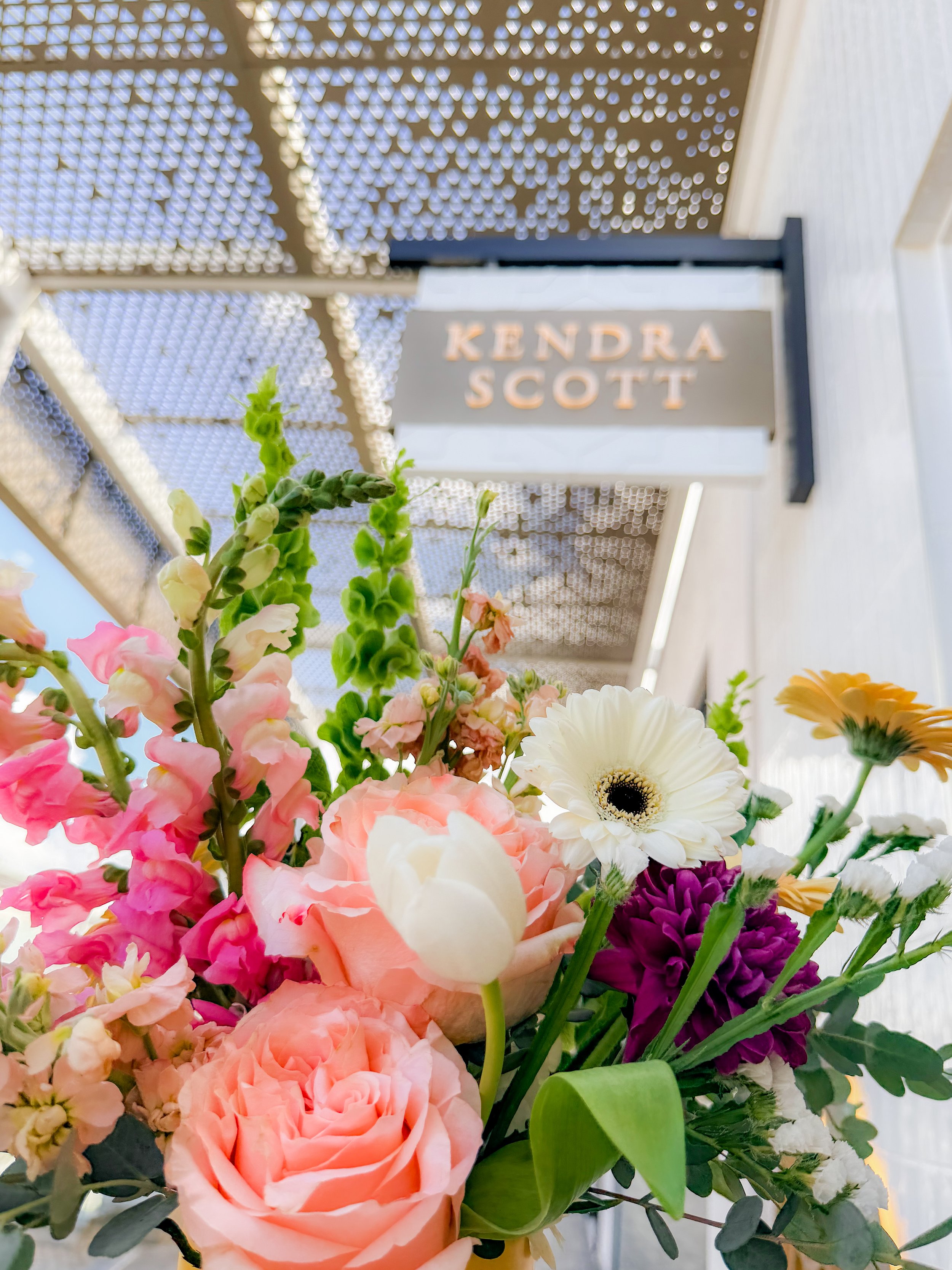 Colorful flower bouquet with pink, white, yellow, purple flowers and green foliage in front of a sign reading 'Kendra Scott' under a decorative metal roof.