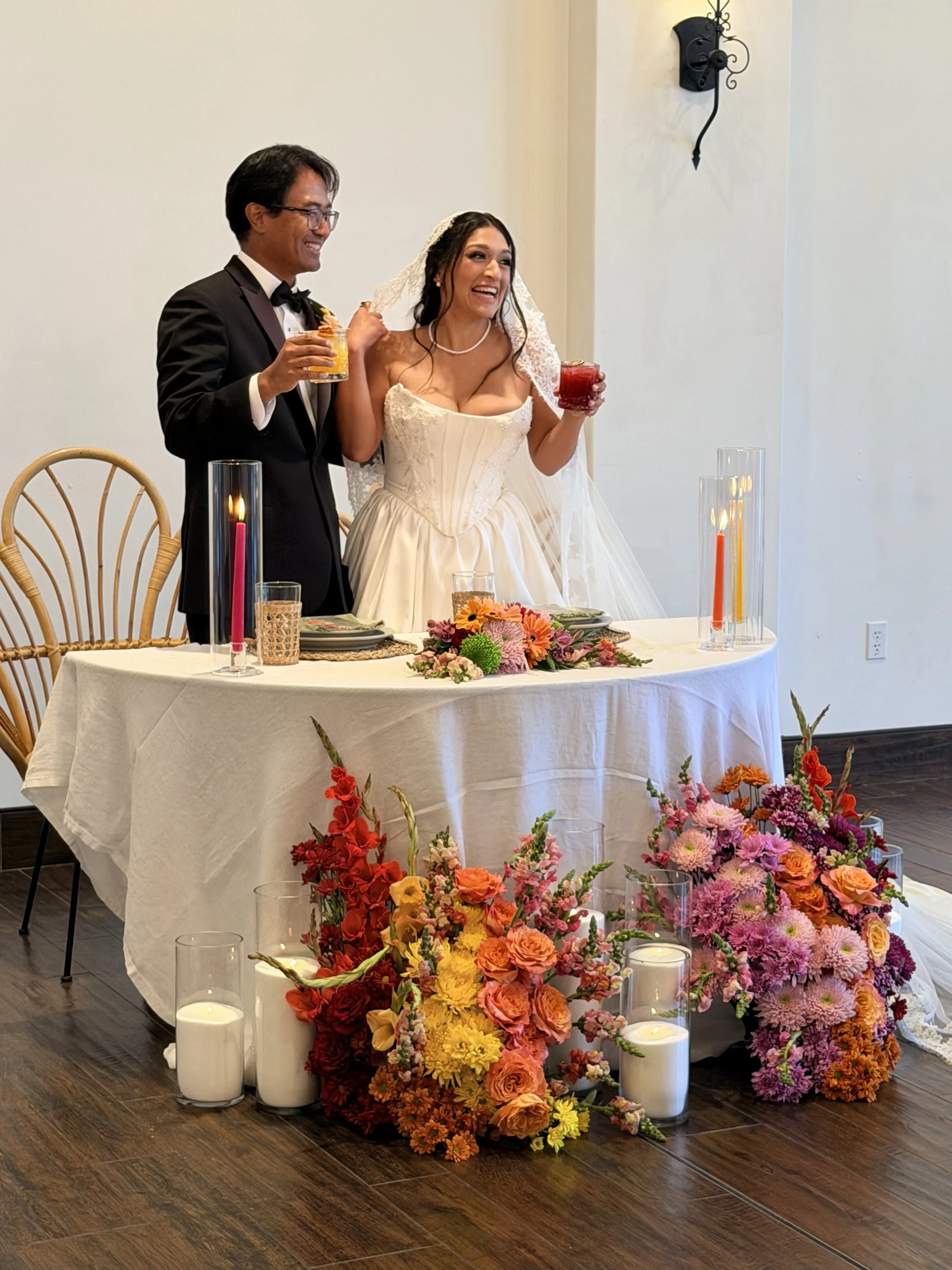 Wedding reception with a bride and groom smiling and holding drinks, surrounded by colorful flowers and candles on a table.