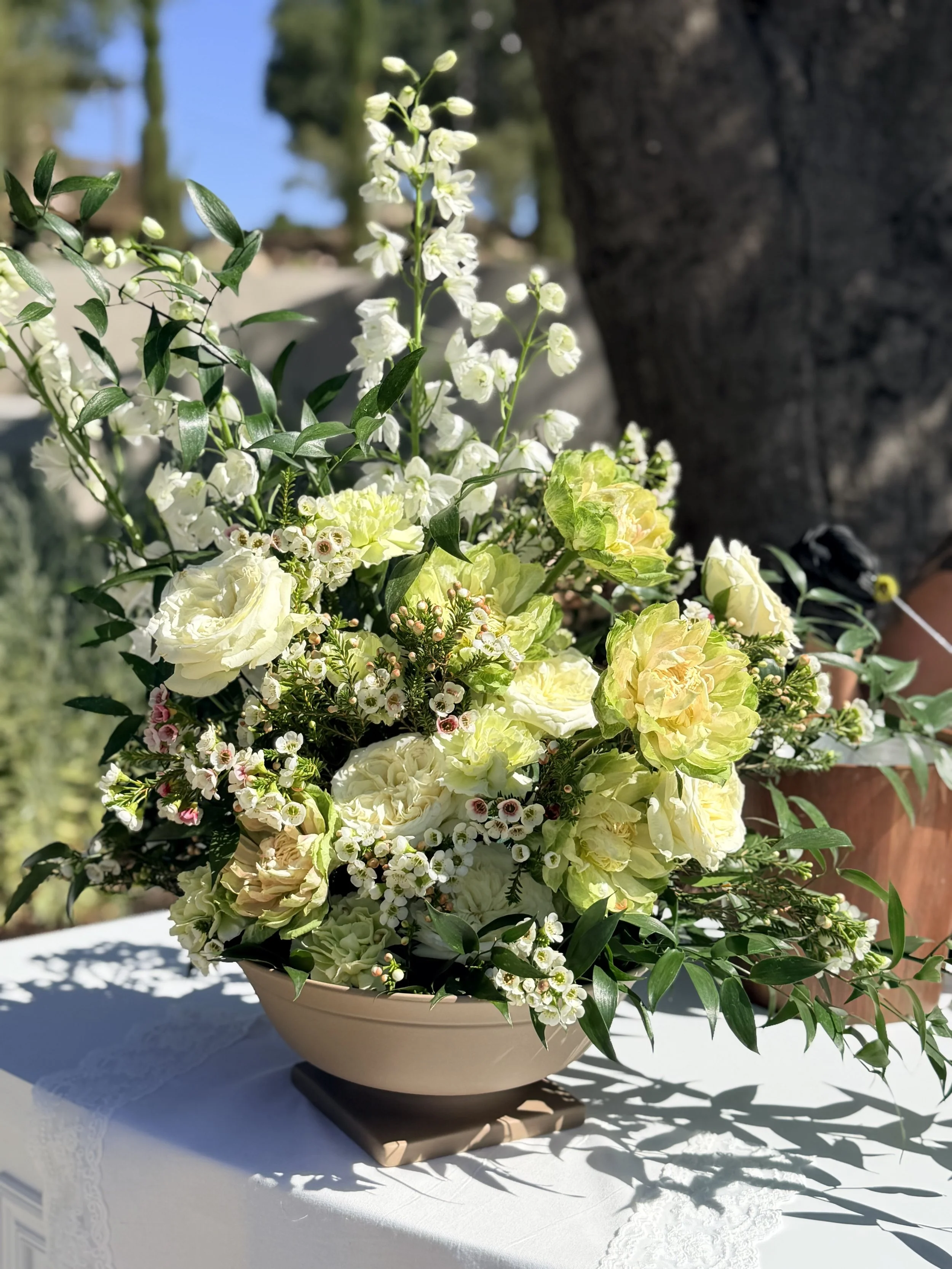 An elegant floral arrangement with white and yellow flowers and green foliage in a beige bowl, placed on a white lace tablecloth outdoors with blurred trees and blue sky in the background.