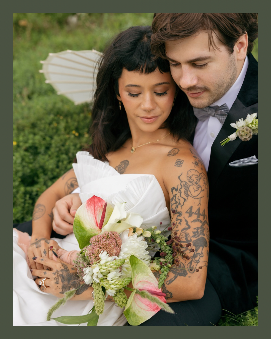 A bride and groom embracing outdoors, holding a bouquet of white and pink flowers, with the bride showing tattoos on her arm.