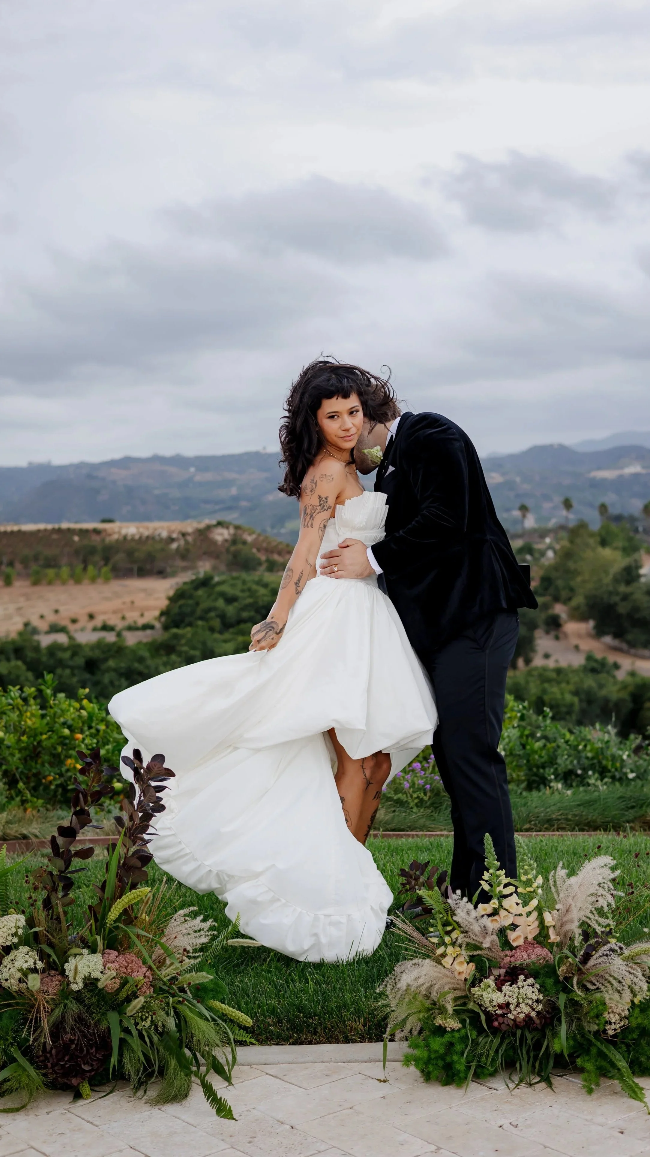 A bride and groom embracing outdoors during a wedding, with scenic hills and cloudy sky in the background, floral arrangements at their feet.