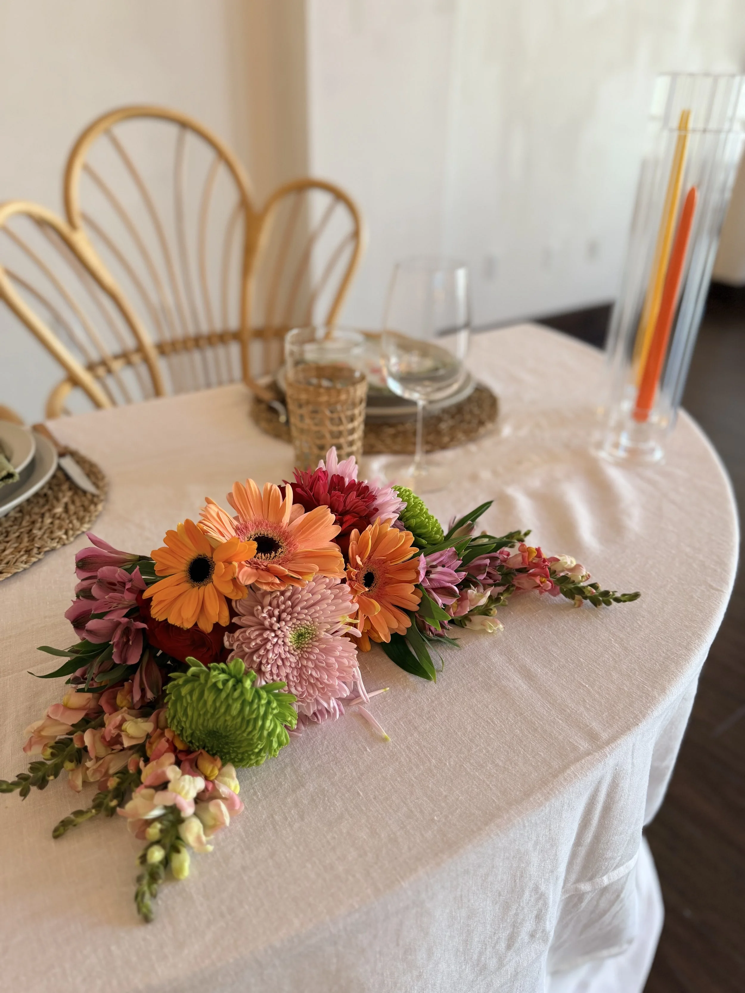 A table with a pink tablecloth features a colorful flower bouquet, glassware, and woven placemats with rattan chairs nearby, in a bright room.
